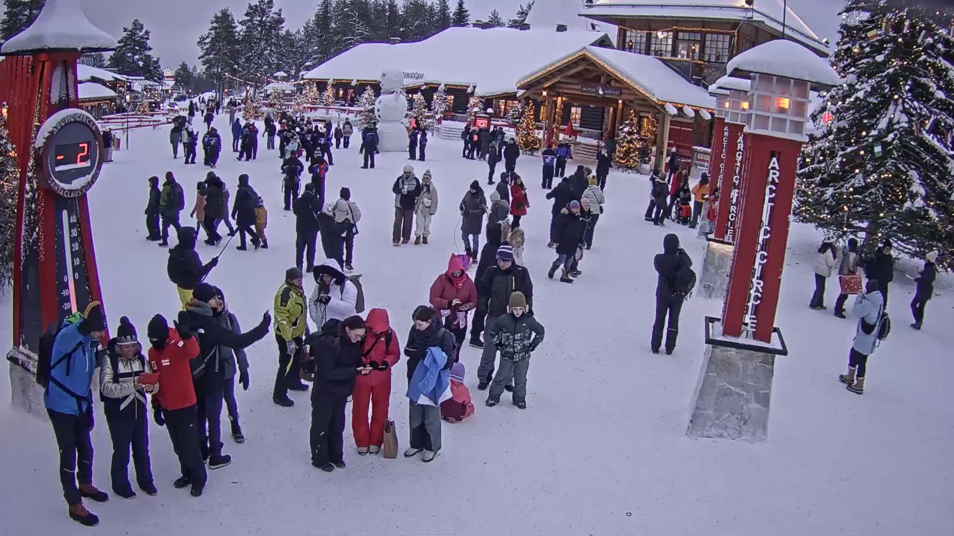 A crowd of people gathers in a snowy plaza in front of a Christmas-themed building, with a large decorated tree and several tall red markers nearby.