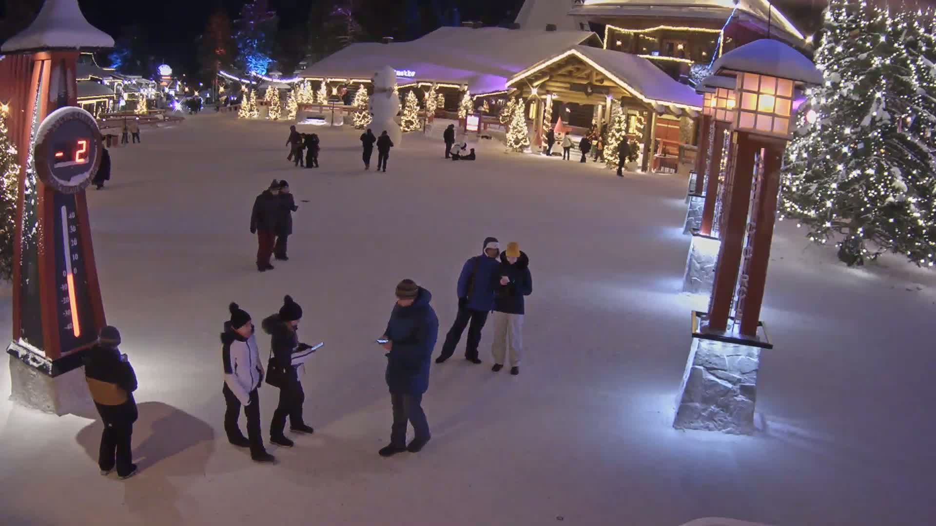 A snow-covered plaza, illuminated at night, features several people standing in small groups, a large thermometer showing -2 degrees, and several decorated Christmas trees near log cabin-style buildings.