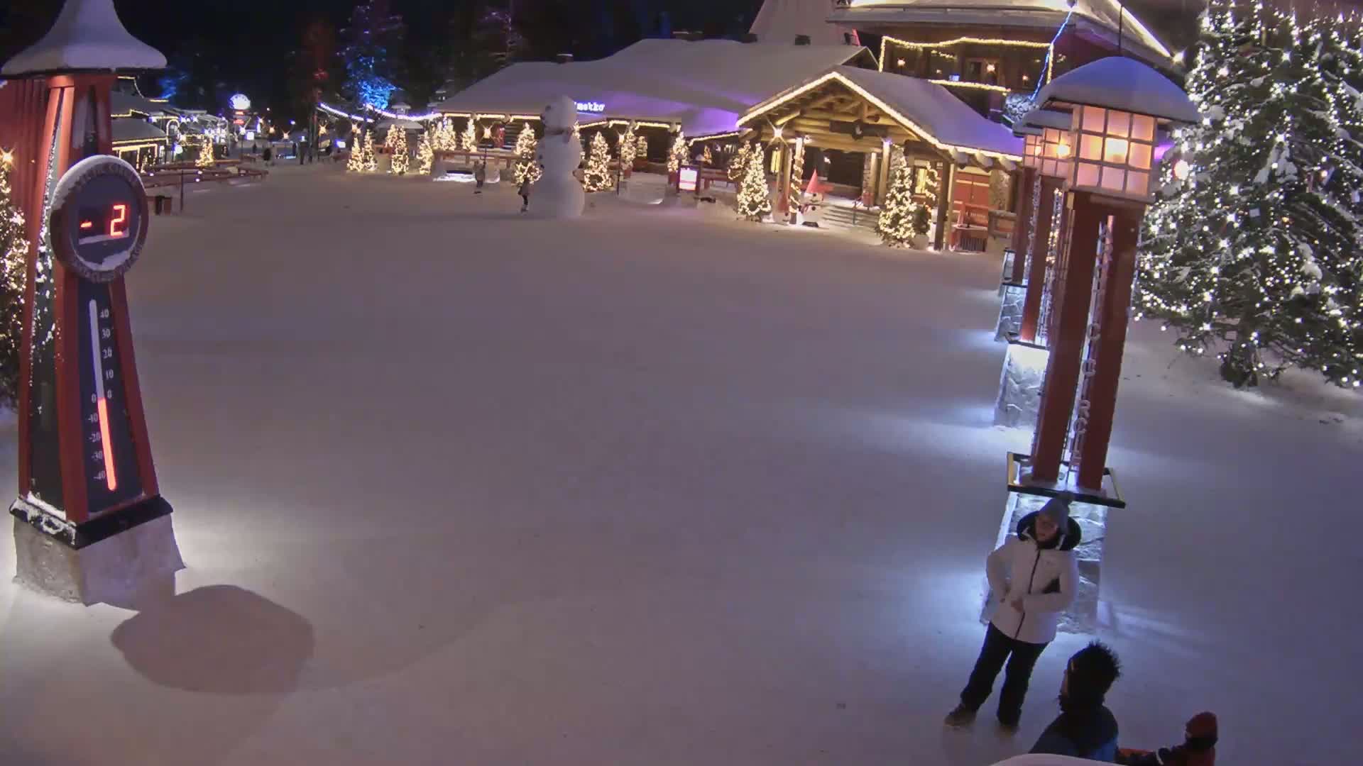 A snow-covered plaza at night, illuminated by lights on buildings and trees, with a thermometer showing -2 degrees and several people standing in the snow.