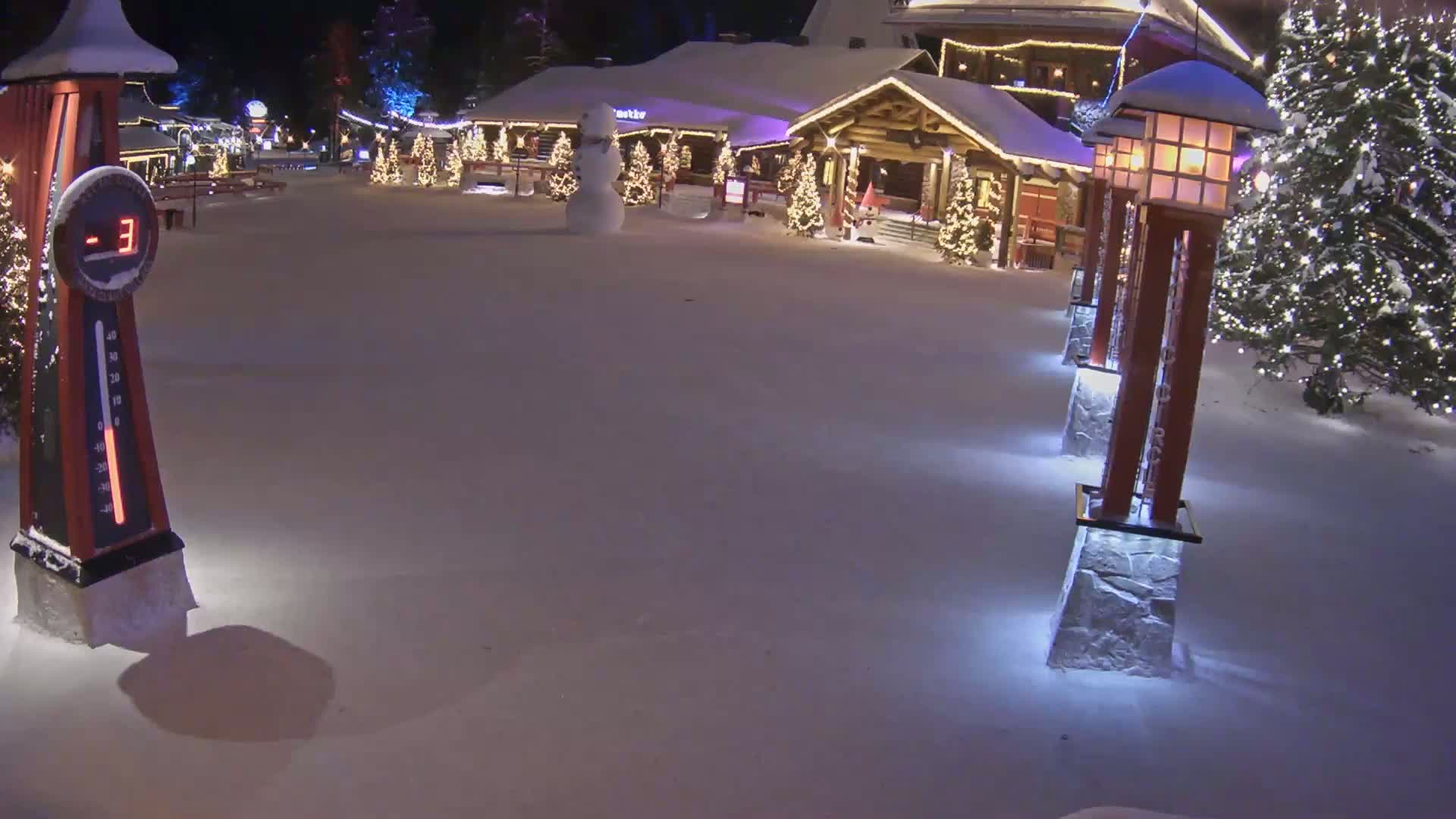 A snow-covered plaza at night features a wooden building decorated with Christmas lights, a snowman, and illuminated lampposts, with the temperature displayed as minus 3 degrees.