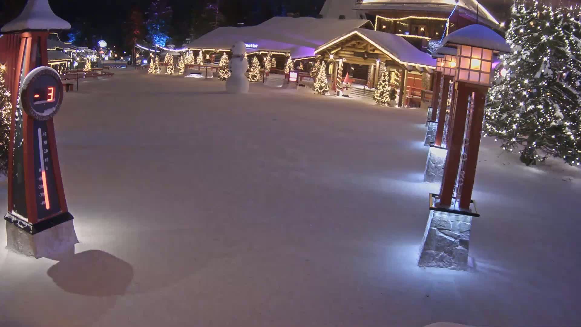 A snow-covered village square at night, illuminated by lights on trees and lampposts, shows a snowman and a thermometer displaying a temperature of -3 degrees.