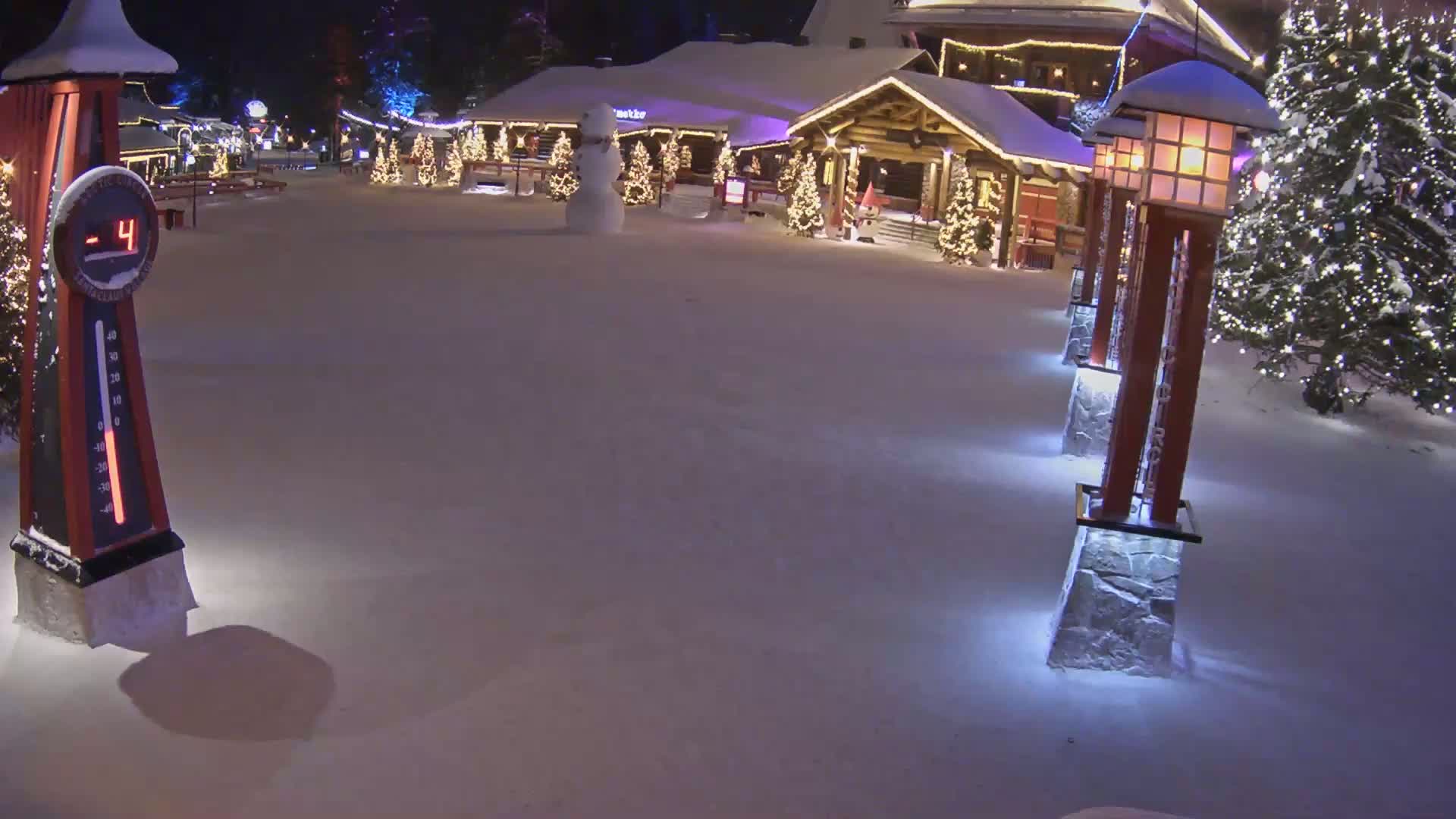 A snow-covered plaza at night, featuring illuminated Christmas trees, a snowman, a wooden building, and decorative light posts, with the temperature showing -4 degrees.