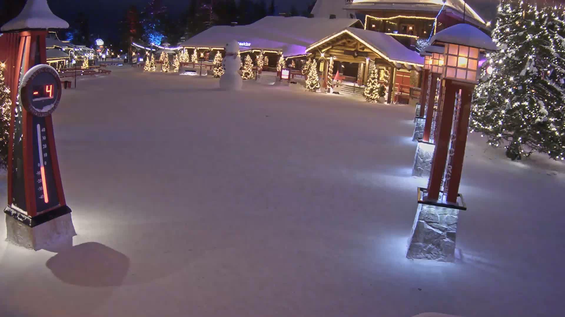 A snow-covered village square at night, illuminated by lights on buildings and lampposts, shows a temperature of -4 degrees, with a snowman in the middle ground.