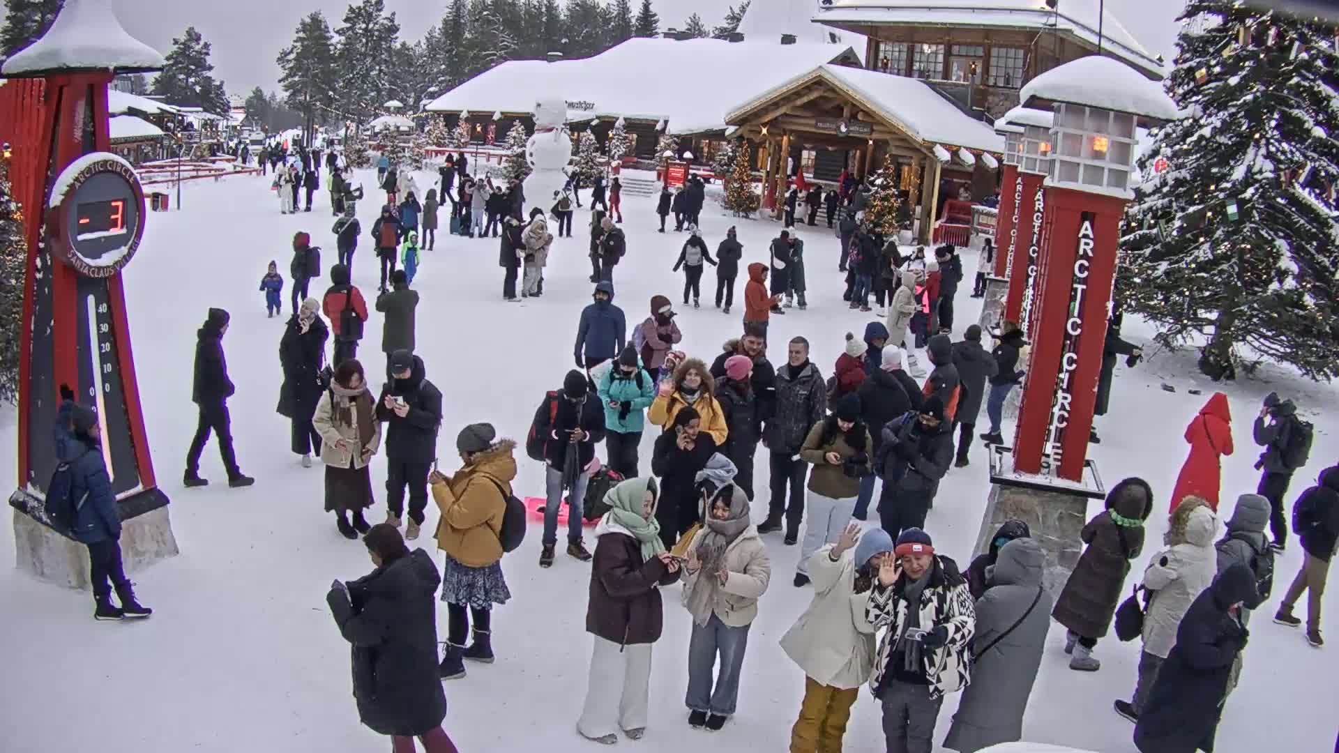 A crowd of people gathers in a snowy plaza in front of wooden buildings, under a gray sky.