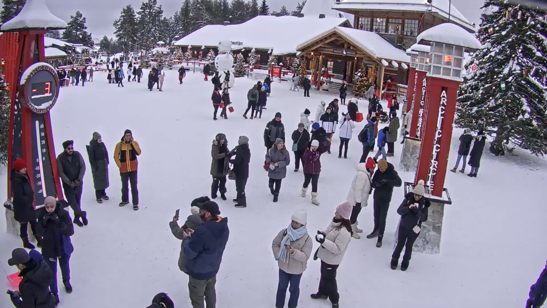 Many people are gathered in a snowy plaza in front of wooden buildings, some taking photos.