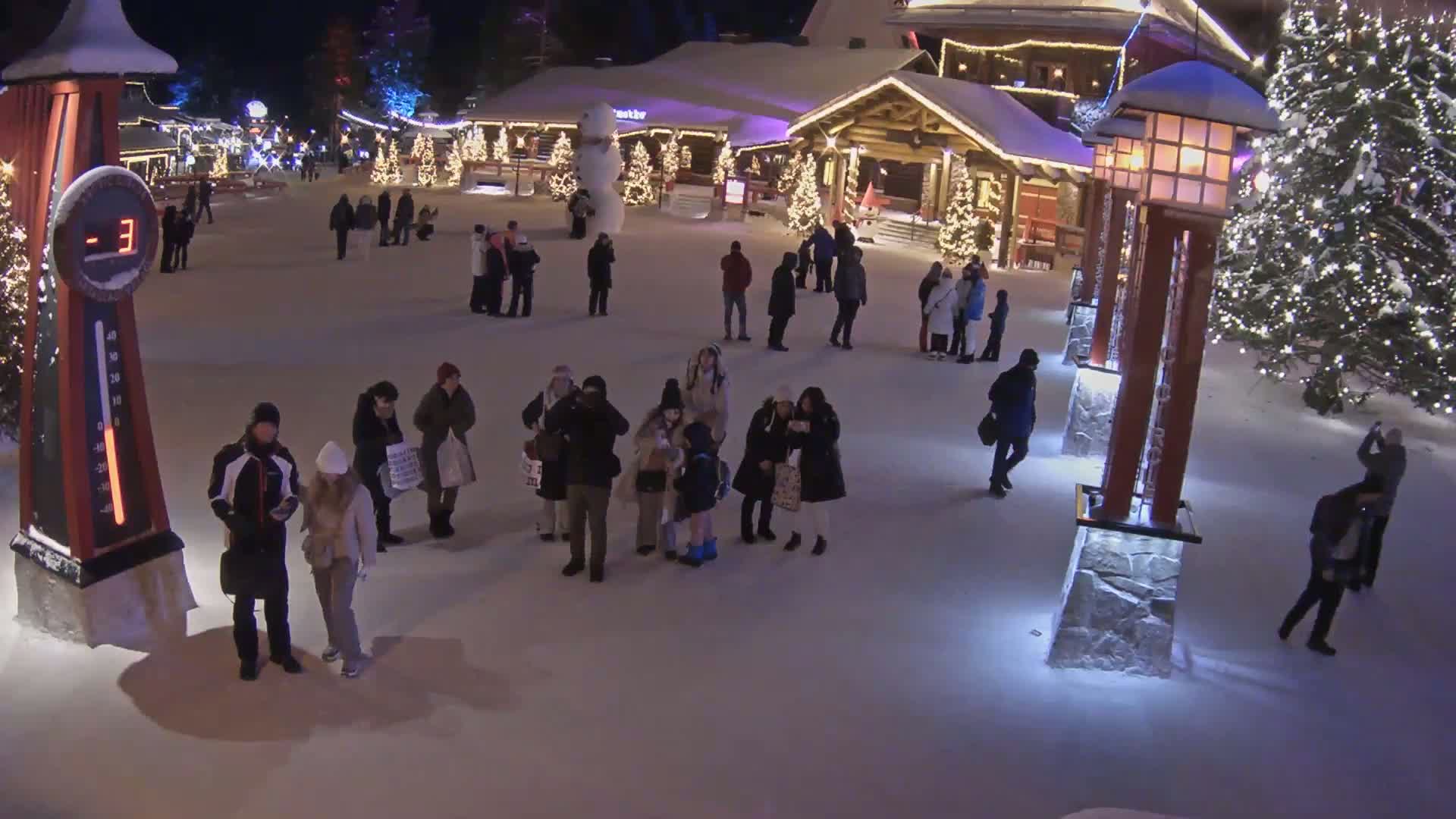 A snow-covered plaza at night, illuminated by lights on buildings and trees, features numerous people walking around in the cold (-3°C).