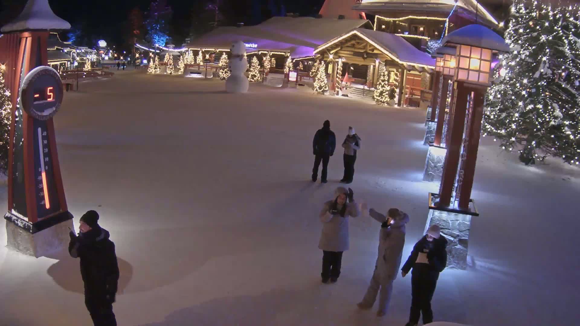 A snow-covered village square at night, illuminated by lights on buildings and lampposts, shows several people walking around, a snowman, and a thermometer displaying -5 degrees.