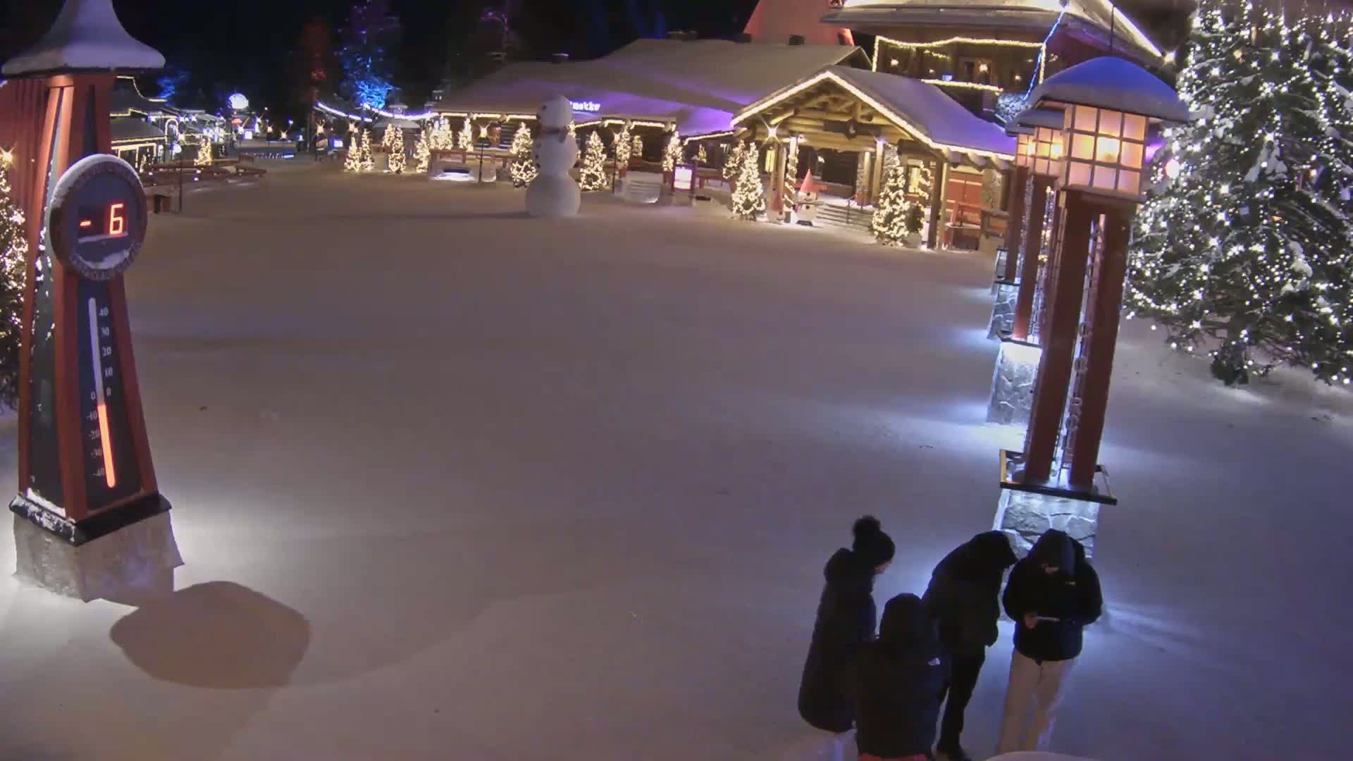 A snow-covered village square at night, illuminated by lights on buildings and trees, shows a group of people standing near a row of lampposts, with a snowman and a thermometer showing -6 degrees visible in the background.