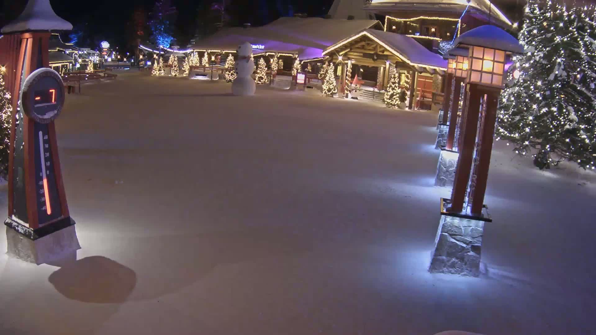 A snow-covered plaza at night features illuminated Christmas trees,  lighted posts, a snowman, and a building with festive decorations, under a clear, dark sky.