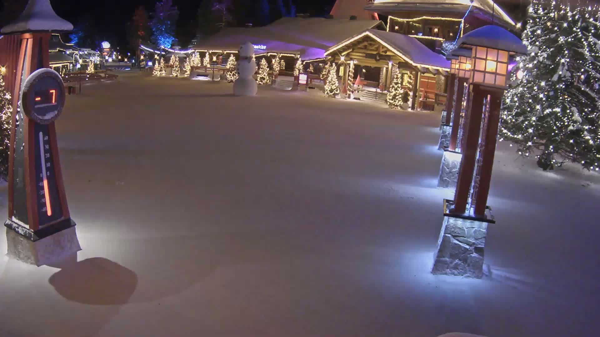 A snow-covered village square at night, illuminated by lights on buildings and lampposts, features a snowman and shows a temperature of -7 degrees.