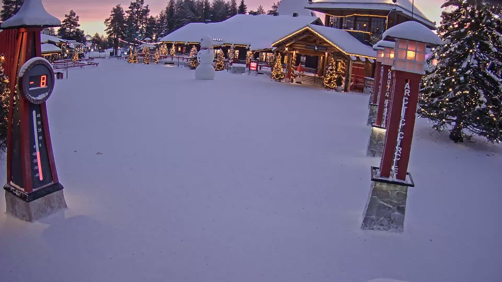 A snow-covered village square at twilight features a building decorated with Christmas lights, a snowman, and a thermometer showing a temperature of -8 degrees.