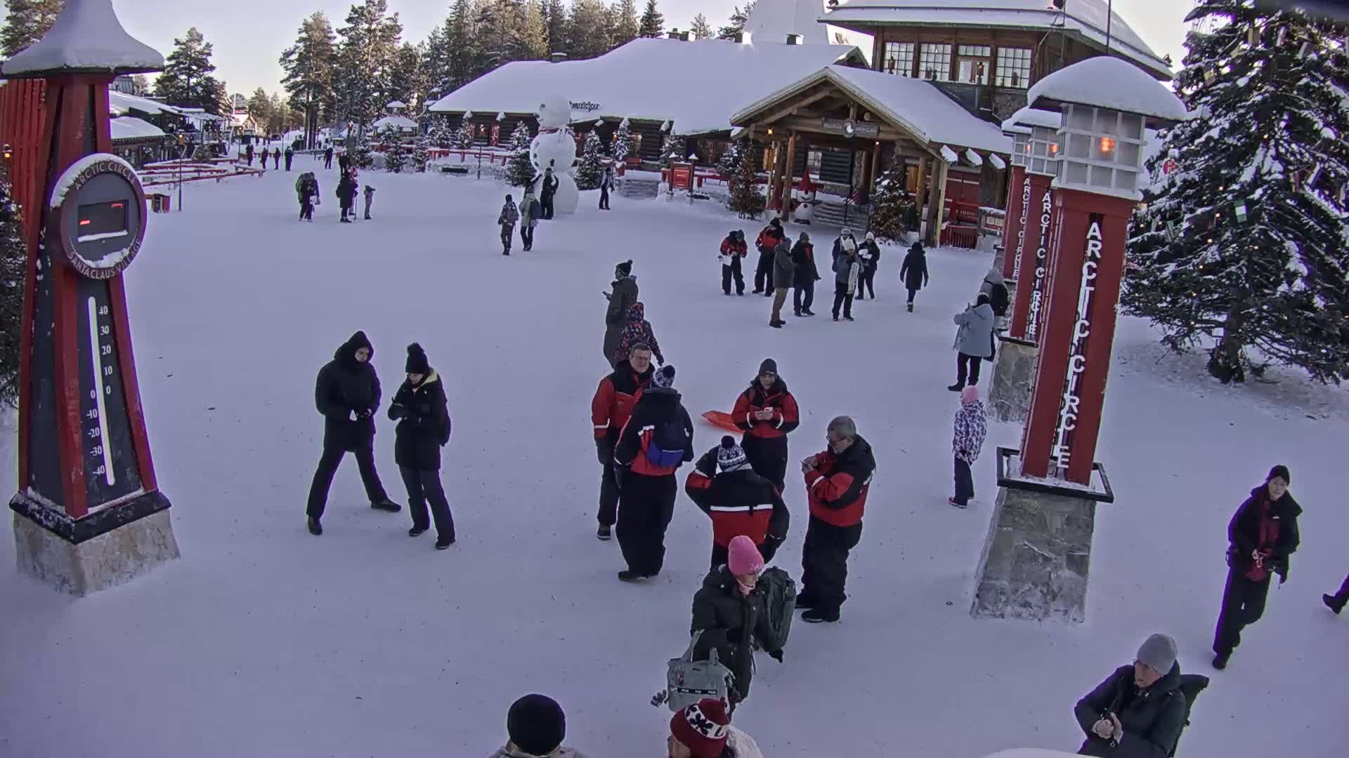 A snowy plaza with several people milling around near a building, a large snowman, and tall red Arctic Circle markers.