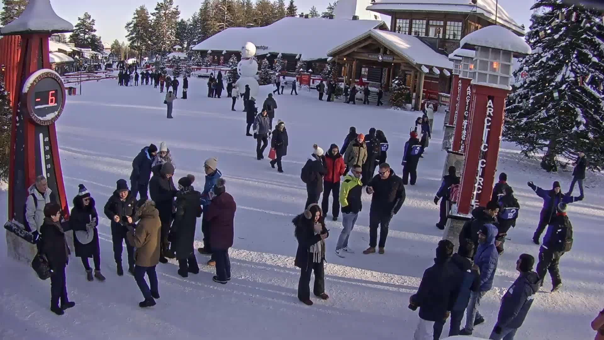 A crowd of people gathers in a snowy plaza on a sunny day, near a building and a snowman, with a thermometer showing a temperature of -6 degrees.