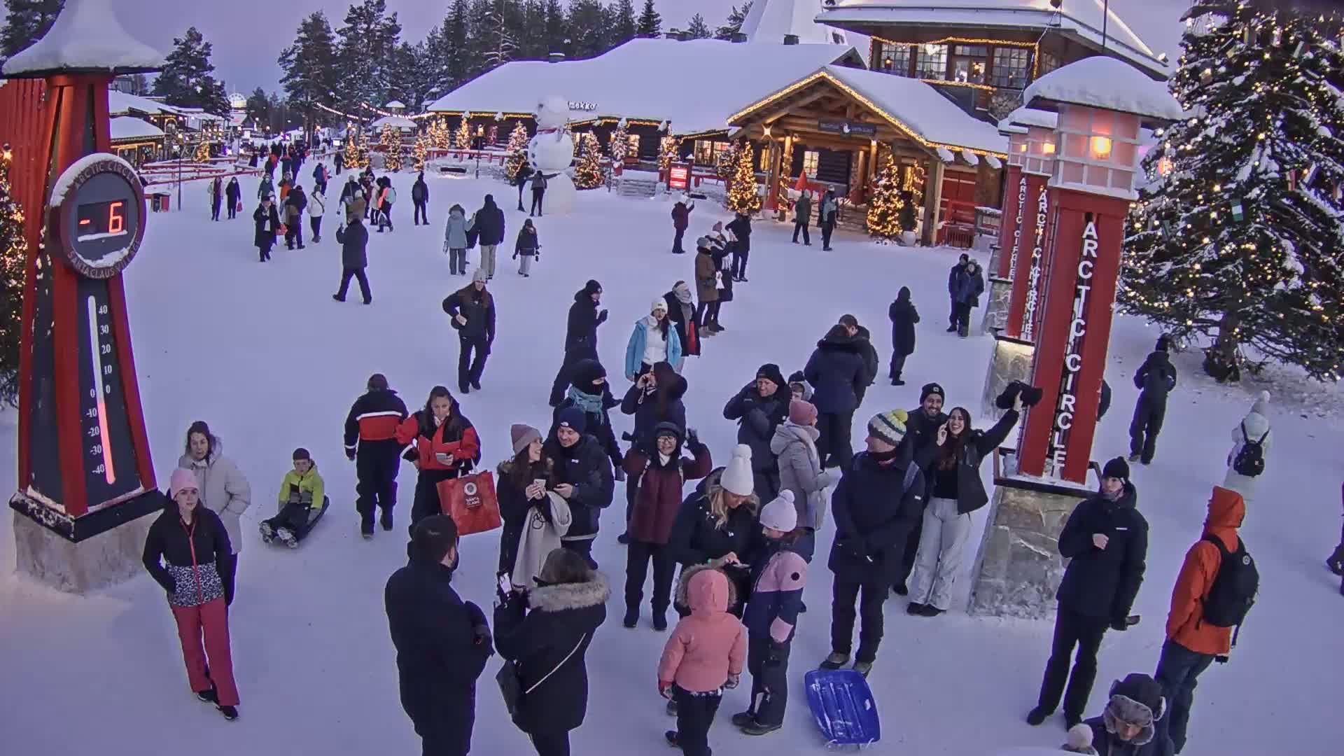 A snowy outdoor plaza with numerous people, some sledding, near festive buildings and a large decorated Christmas tree, under a twilight sky.