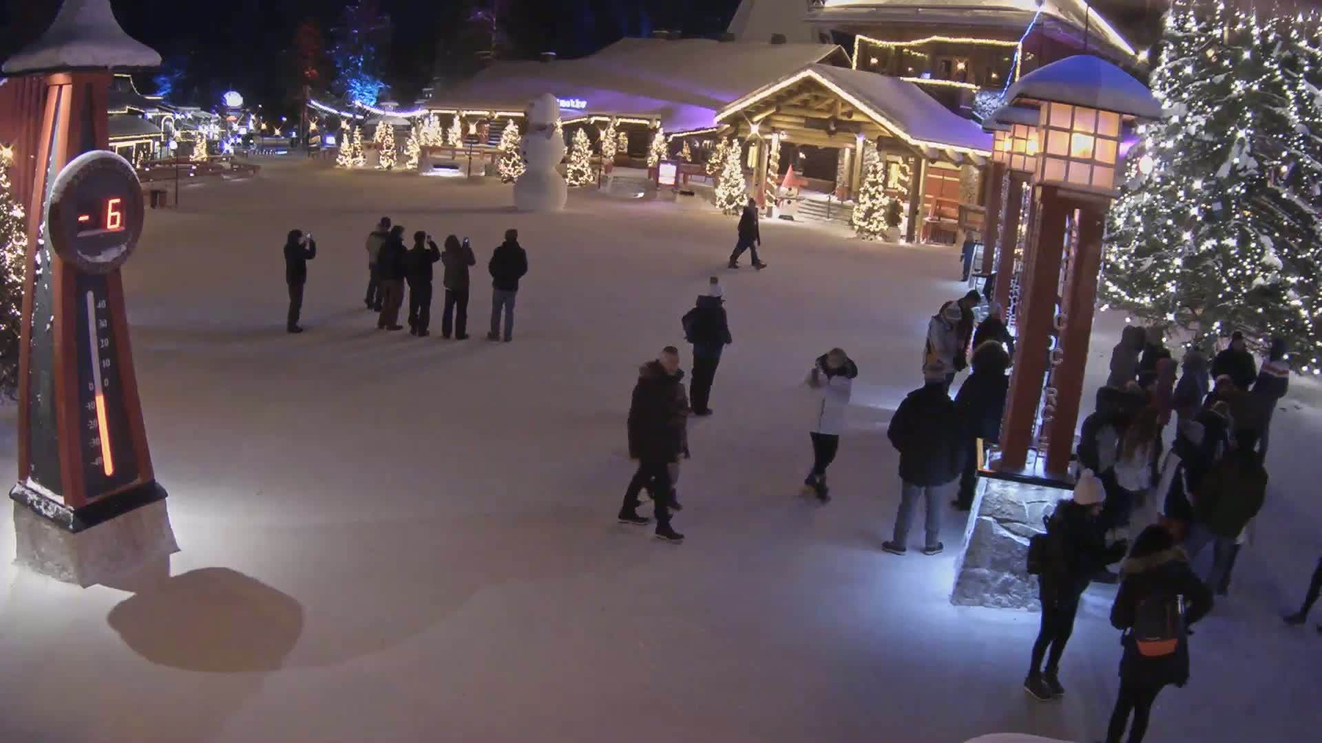 A snow-covered village square at night, illuminated by lights on buildings and trees, shows several people walking around in winter clothing, with a large snowman and a thermometer displaying -6 degrees.