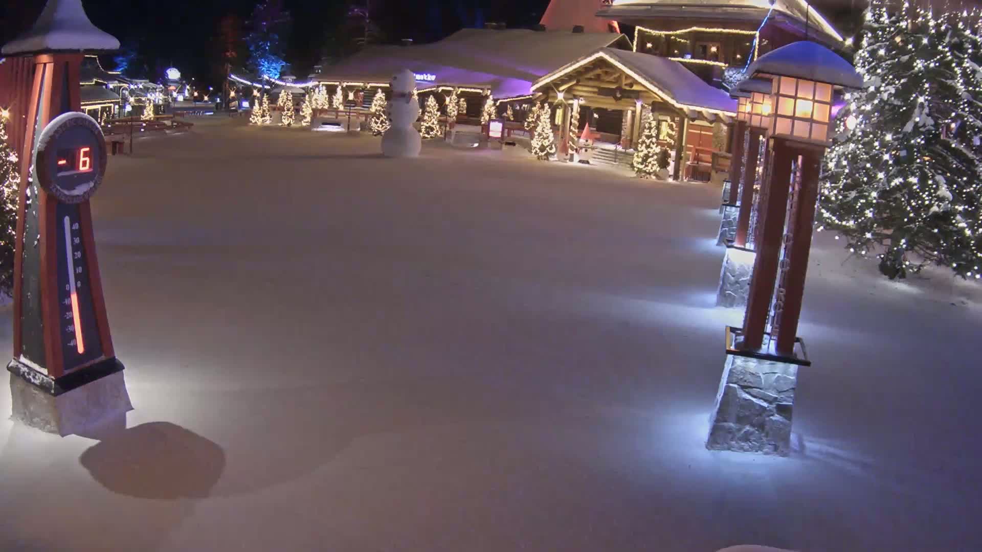 A snow-covered plaza at night, featuring a snowman, illuminated Christmas trees, and lit lampposts, with a thermometer indicating a temperature of -6 degrees.