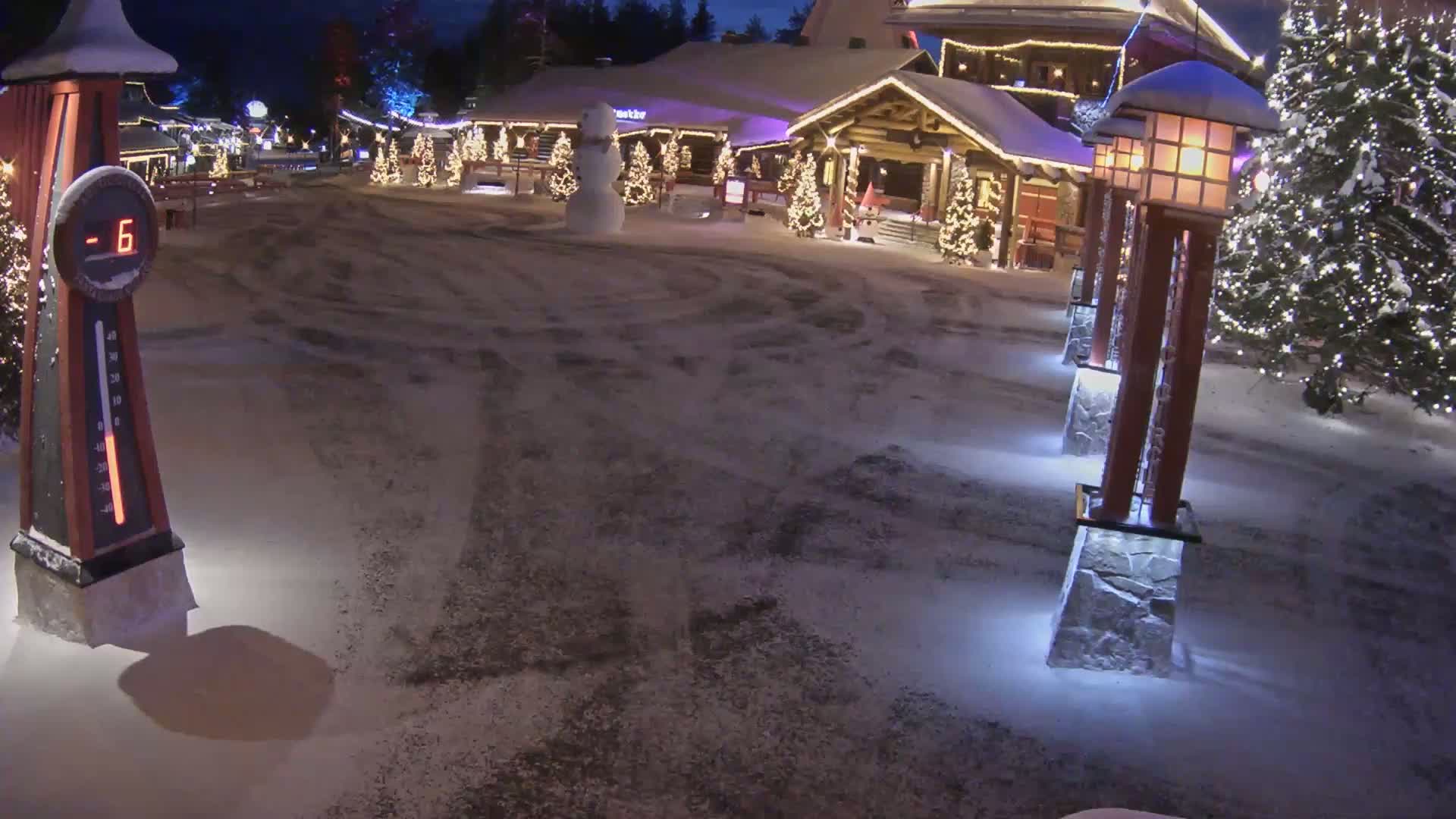 A snow-covered plaza at night features a snowman, illuminated Christmas trees, and a wooden building, with the temperature showing -6 degrees.