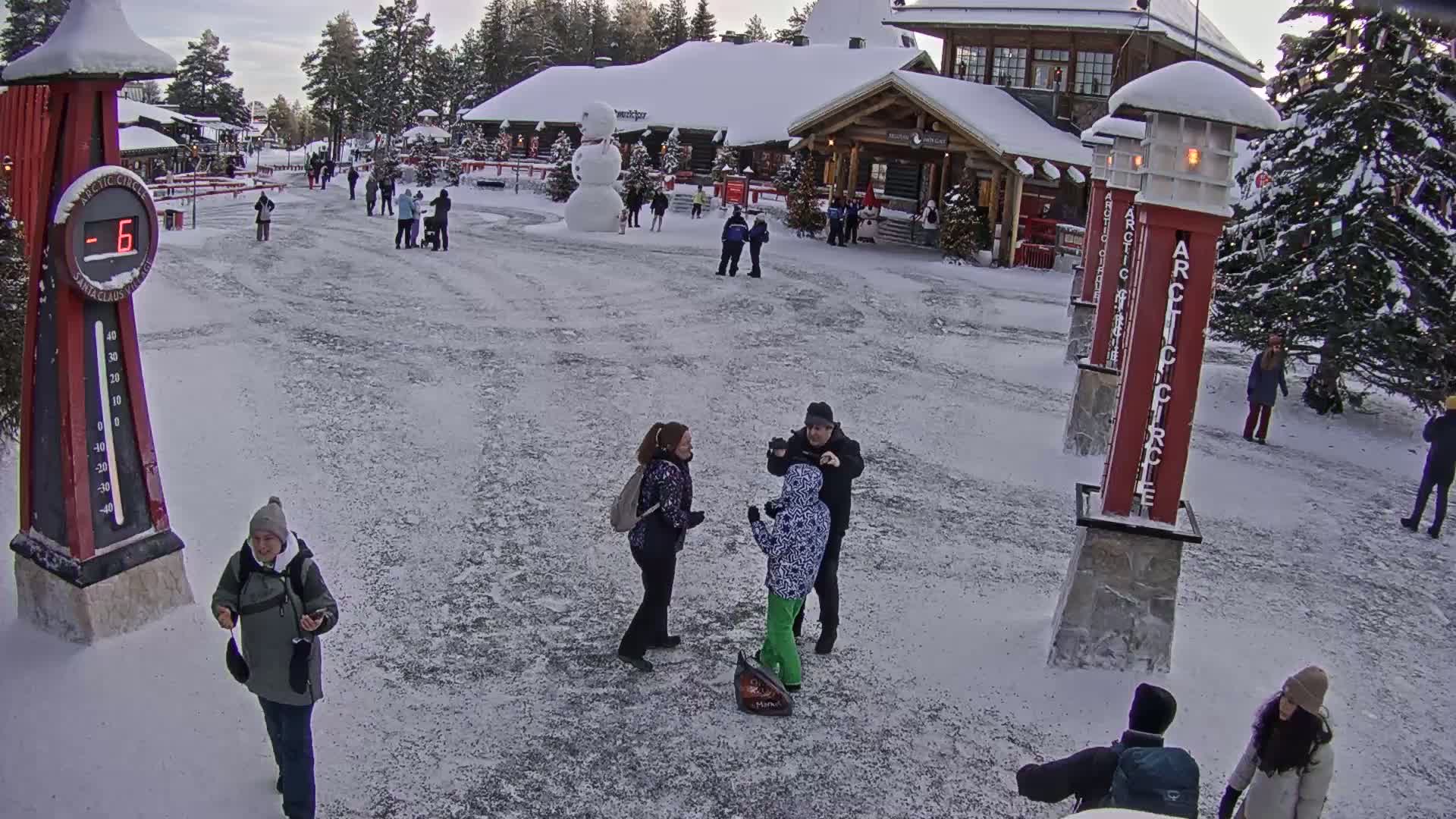 A snow-covered plaza with several people, a large snowman, and wooden buildings in the background, under a cloudy sky.
