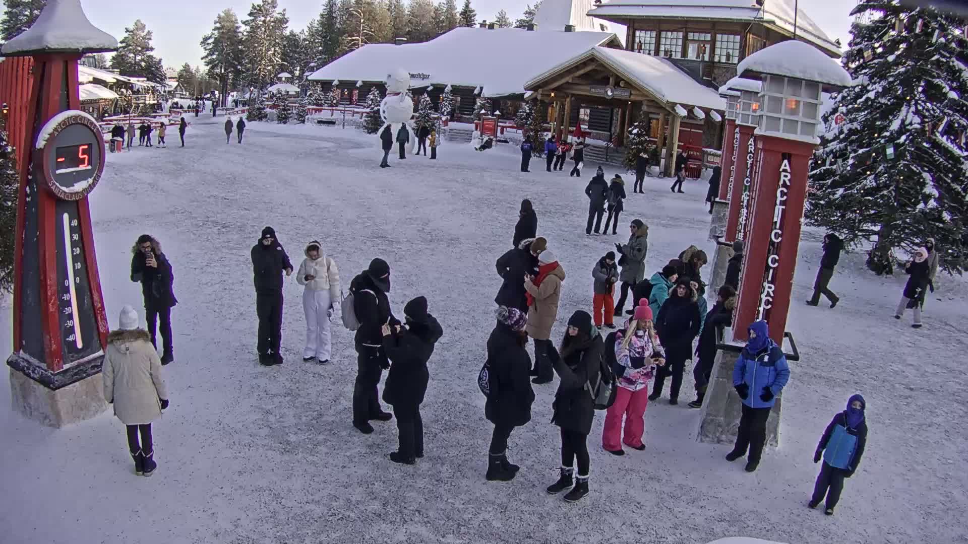 A snowy outdoor plaza features numerous people gathered around a large thermometer displaying -5 degrees, near a wooden building and a snowman.