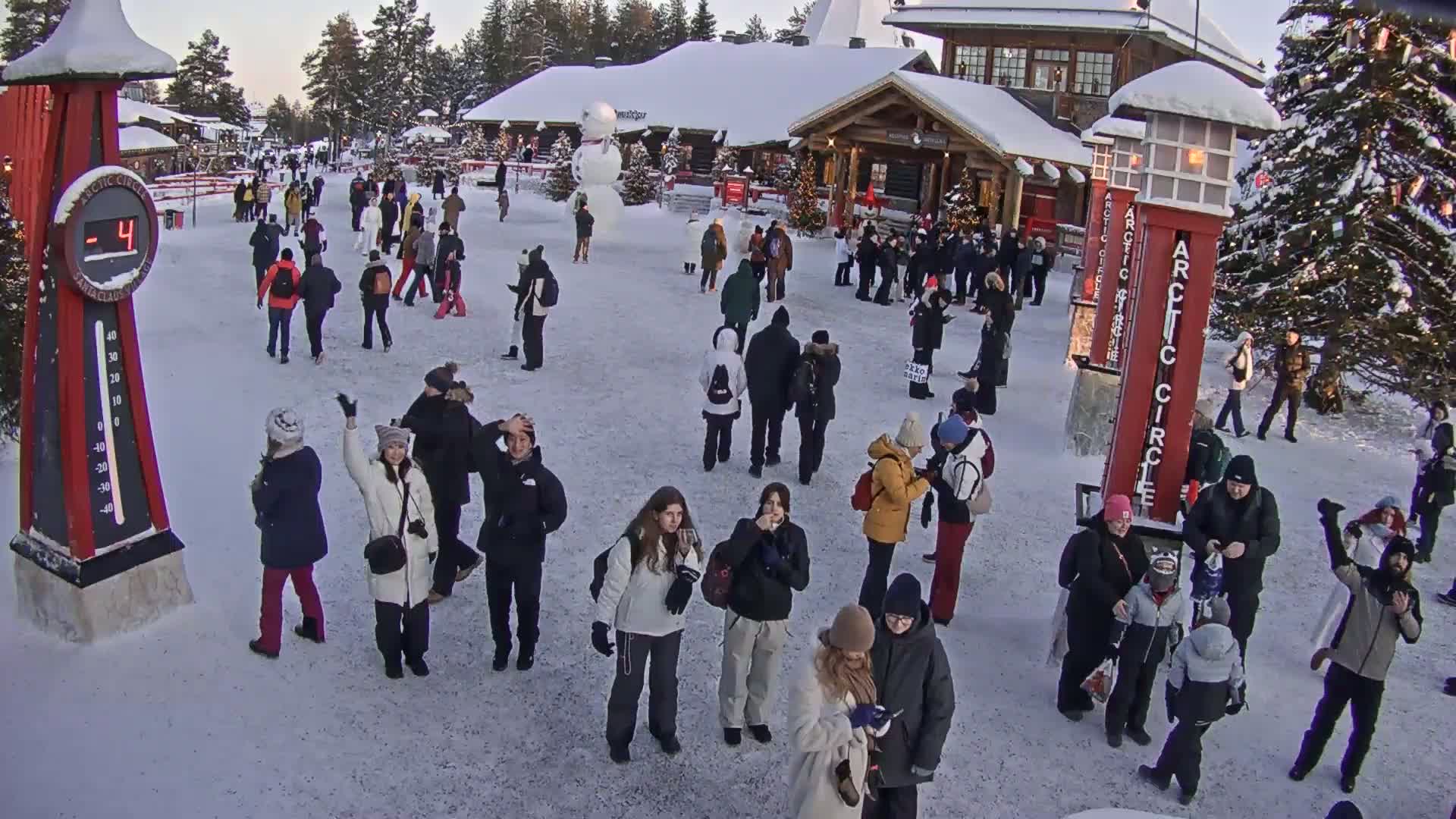 A crowd of people gathers in a snowy plaza, near a large snowman and a building, under a clear twilight sky, with the temperature at -4 degrees.
