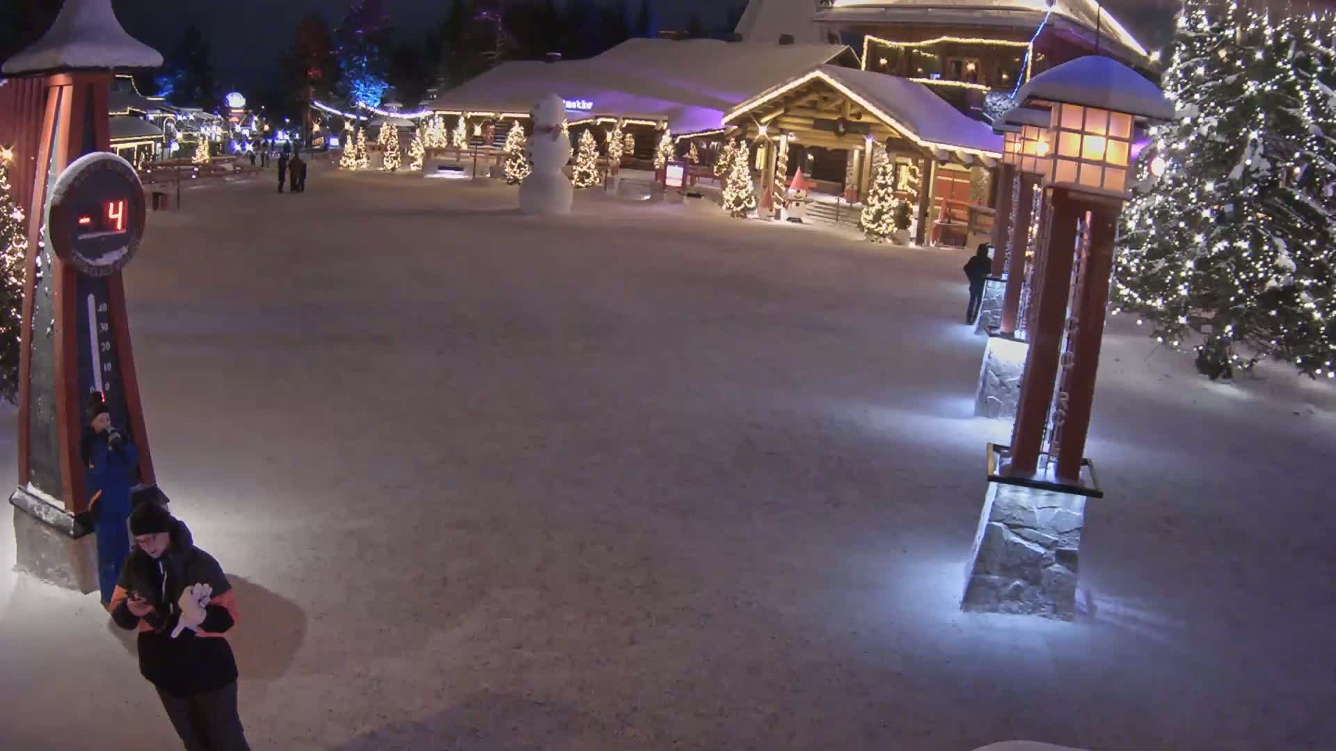 A snow-covered village square at night, illuminated by lights on buildings and lampposts, with a snowman and several people present, and a temperature of -4 degrees.