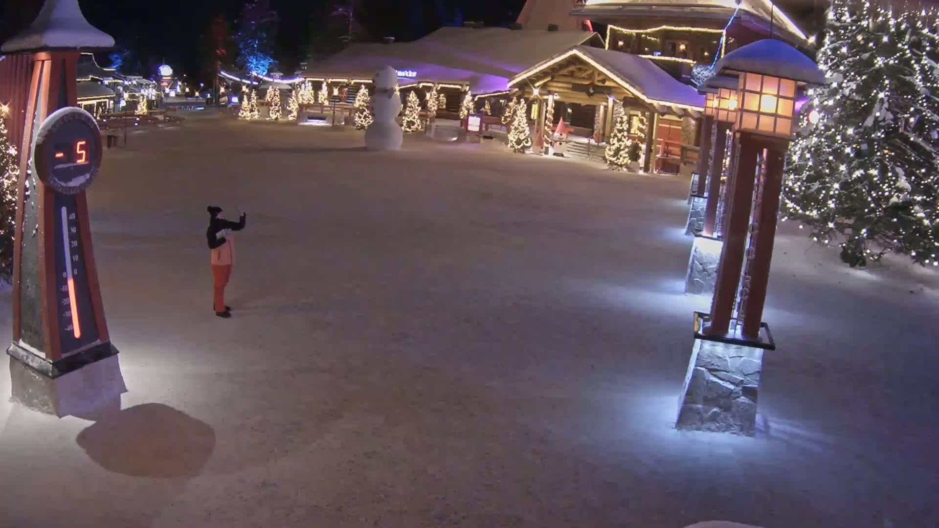 A person takes a picture in a snow-covered plaza at night, illuminated by lights on buildings and lampposts, with a snowman and Christmas trees visible.
