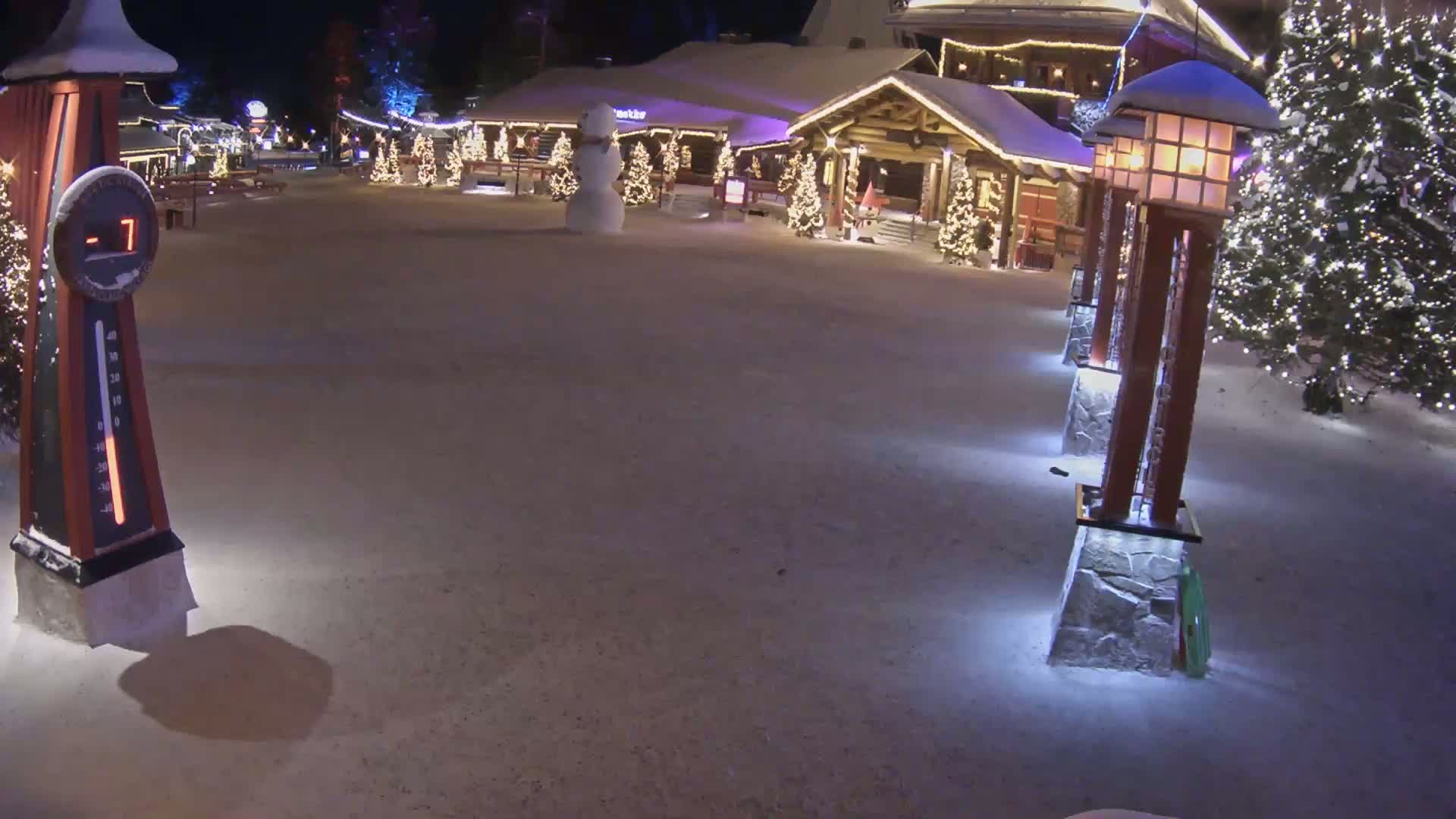A snow-covered plaza at night features a snowman, illuminated Christmas trees, and a series of lit lanterns, with a thermometer displaying -7 degrees.