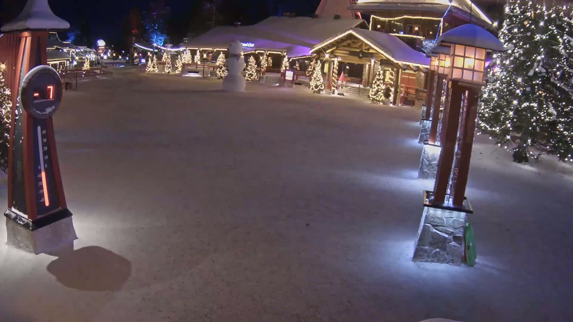 A snow-covered plaza at night, illuminated by lights on wooden posts, features a snowman near a festively decorated building, with the temperature shown as minus seven degrees.