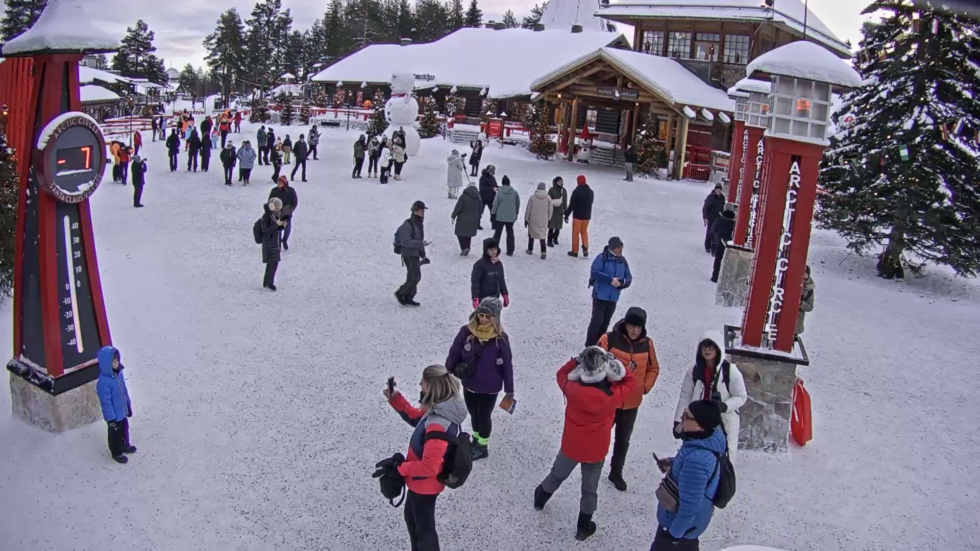 A snowy plaza with numerous people milling about near wooden buildings, a snowman, and a large thermometer displaying minus seven degrees.
