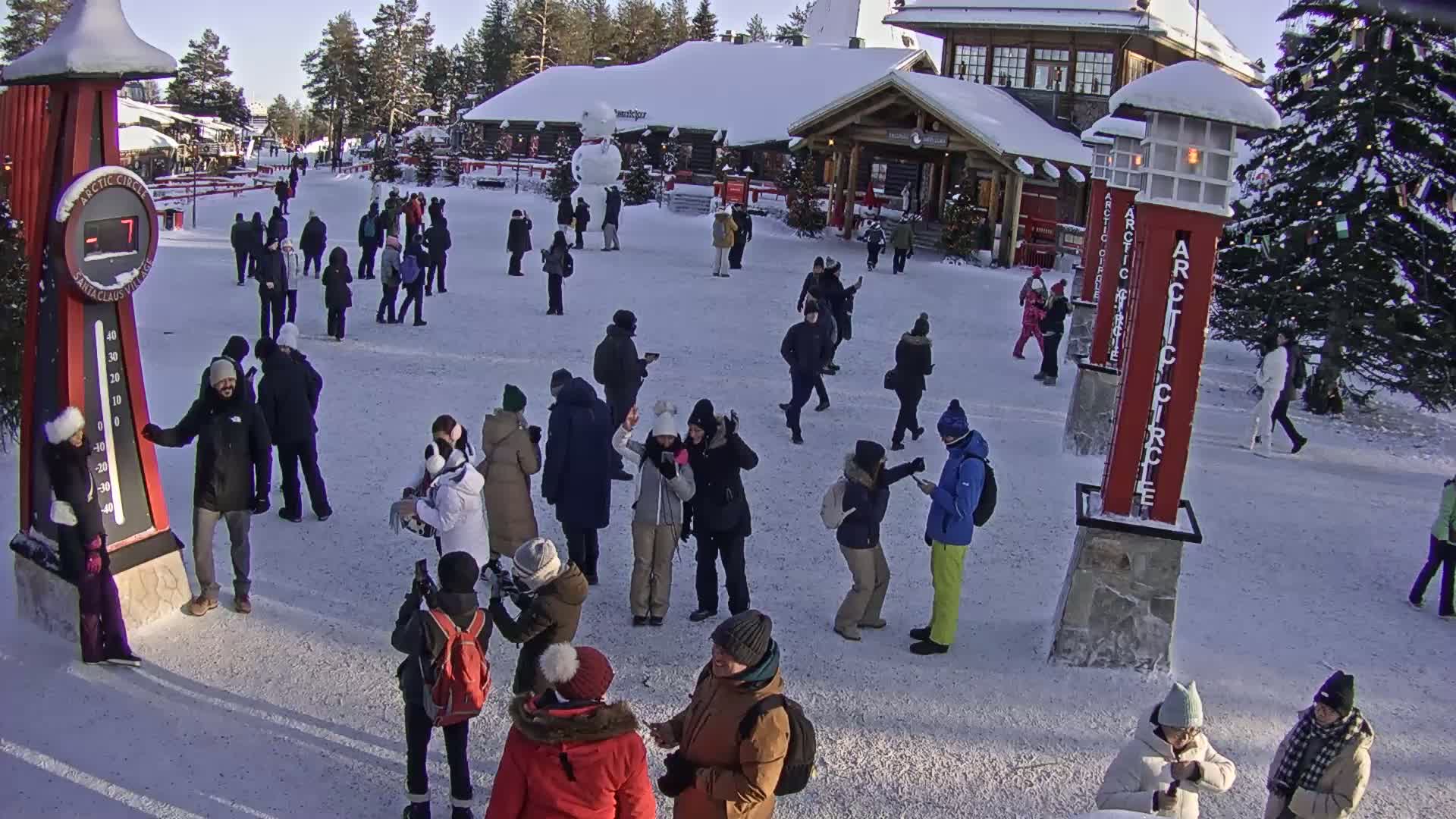 A snowy plaza with many people gathered around, near buildings and a large snowman, on a sunny day.