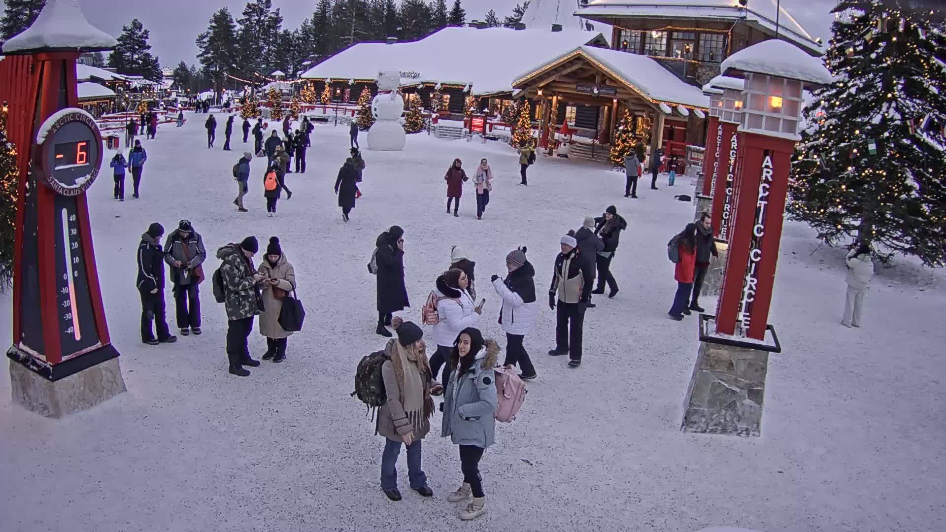 A snowy plaza with several people walking around near wooden buildings and Christmas trees under a twilight sky.