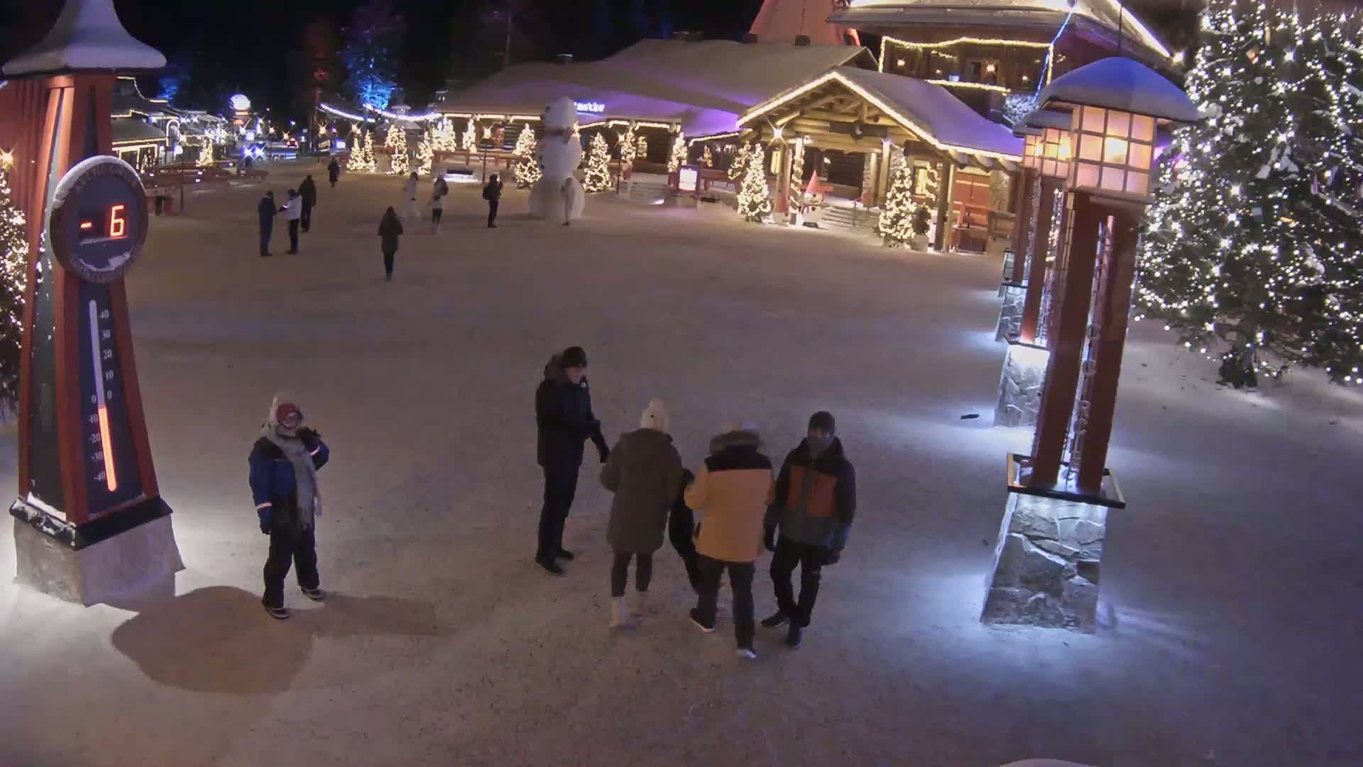 A snow-covered village square at night, illuminated by lights on buildings and lampposts, with several people standing in small groups and a large snowman visible in the background, at -6 degrees Celsius.