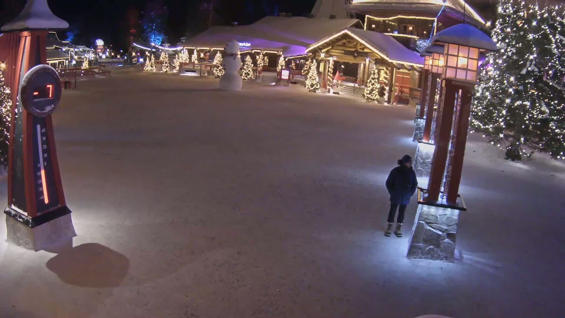 A snow-covered plaza at night, illuminated by lights on buildings and lampposts, features a snowman, decorated Christmas trees, and a person standing near a large thermometer showing a temperature of -7 degrees.