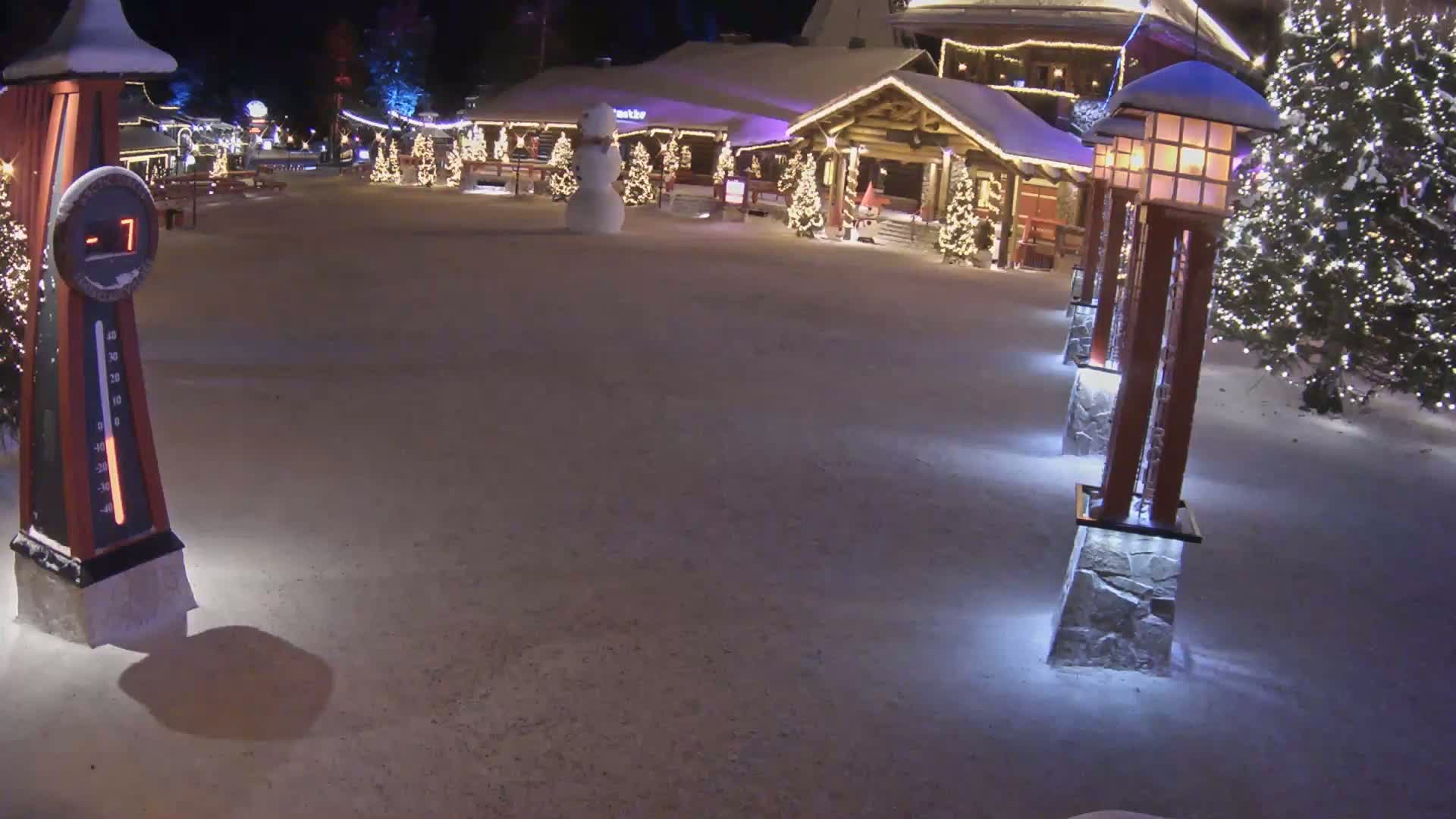 A snow-covered plaza at night features illuminated Christmas trees, a snowman, and several decorative light posts, with a thermometer indicating a temperature of -7 degrees.