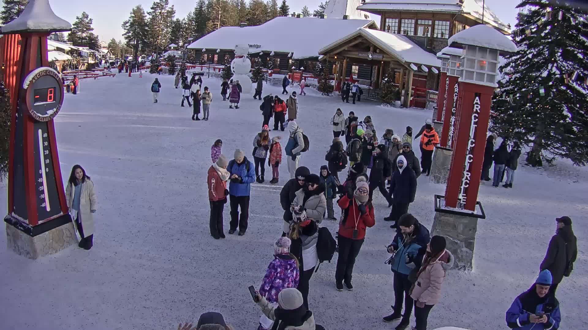 A crowd of people gathers in a snowy plaza in front of wooden buildings under a sunny winter sky.