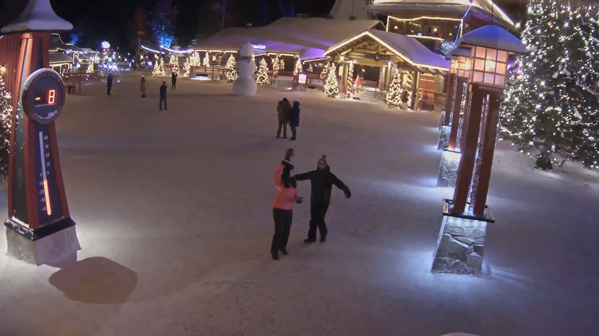A snow-covered village square at night, illuminated by lights on buildings and lampposts, with several people walking around, and a snowman standing near a building.