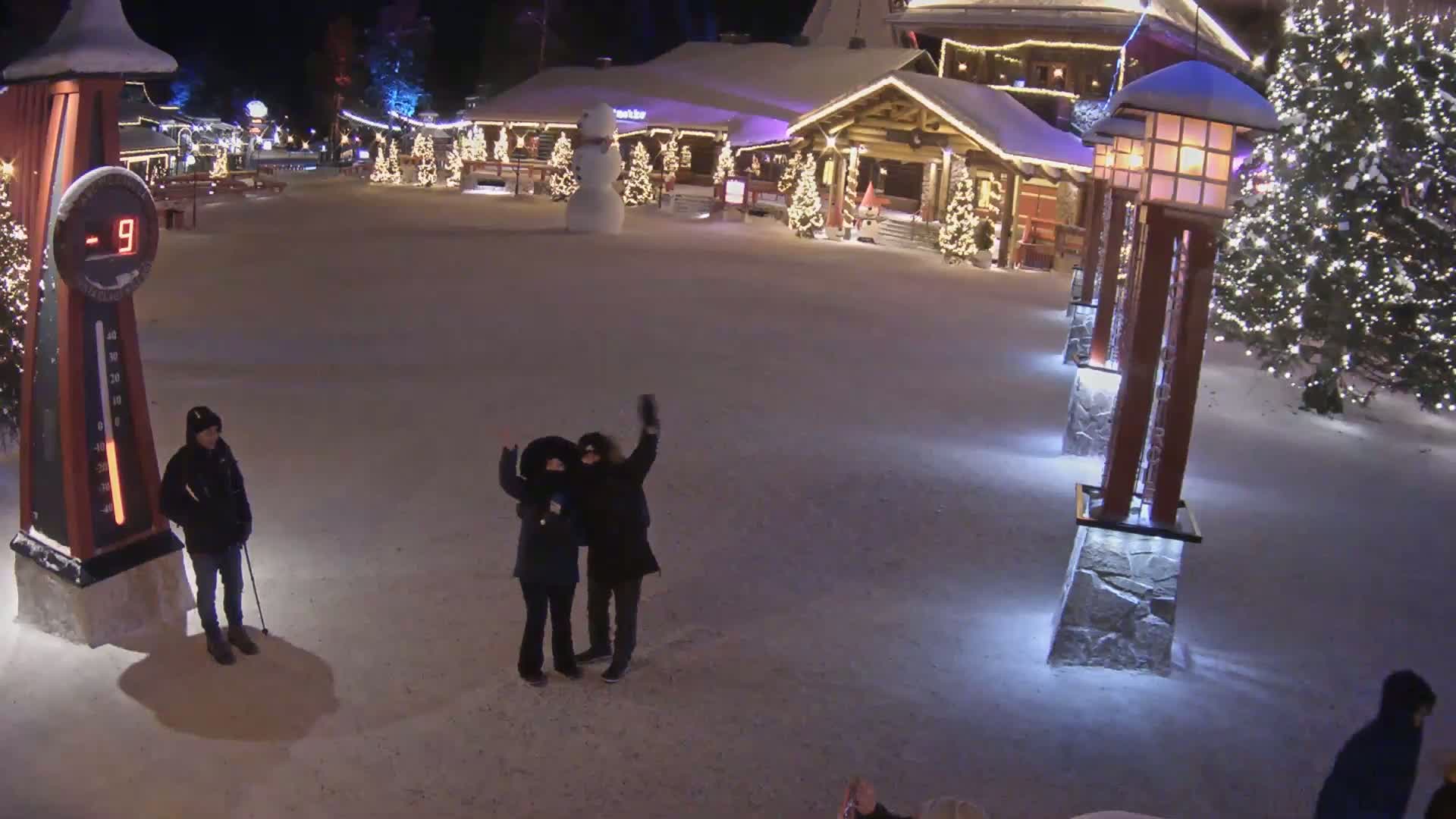 A snow-covered village square at night, illuminated by lights on buildings and lampposts, features a large snowman, several people, and a thermometer showing -9 degrees.