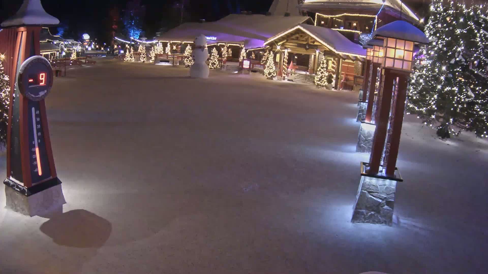 A snow-covered village square at night, illuminated by lights on buildings and lampposts, shows a snowman and a thermometer displaying -9 degrees.