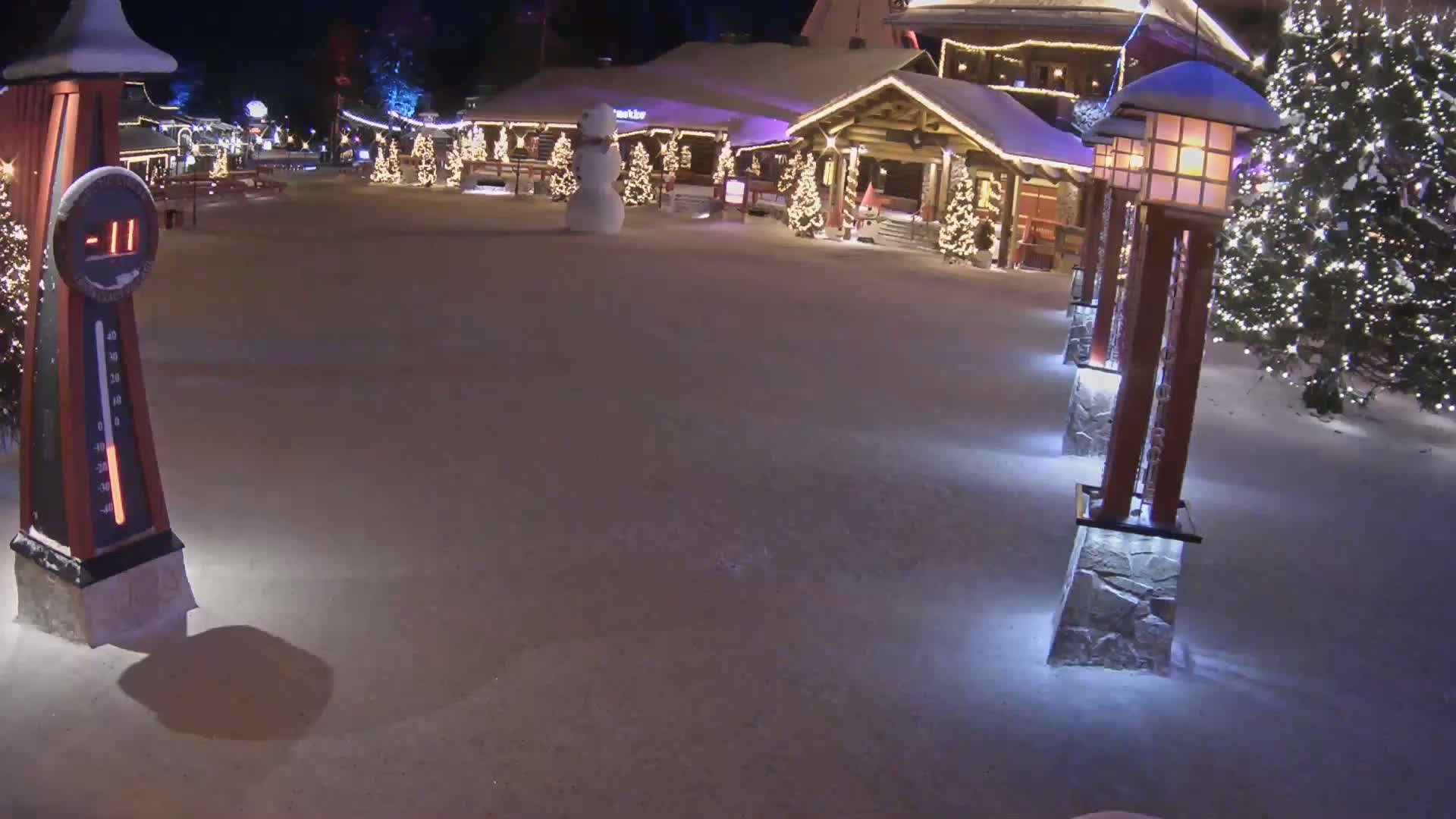 A snow-covered plaza at night, illuminated by lights on lampposts and Christmas trees, features a snowman and shows a temperature of -11 degrees.