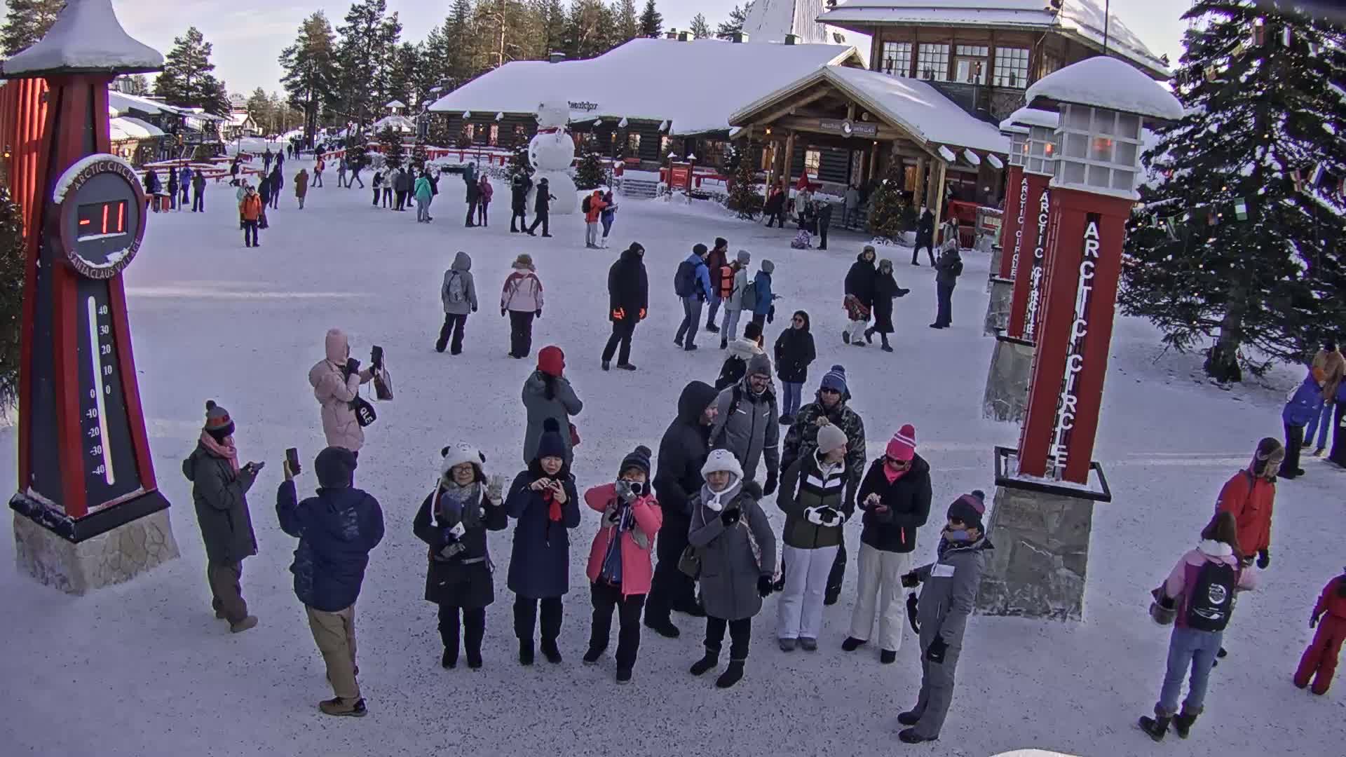 A snowy outdoor scene shows a crowd of people gathered in a plaza in front of a building, with several tall red markers and a large thermometer.