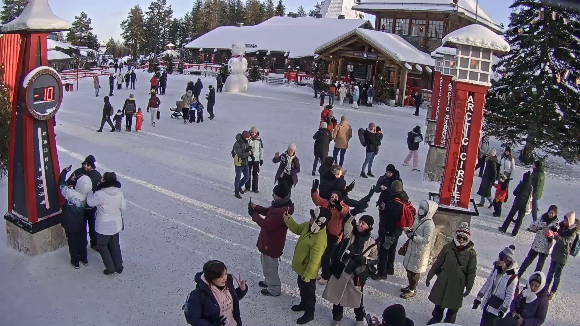 A snowy, outdoor plaza filled with many people, some taking photos, near buildings and a snowman, with a thermometer showing -10 degrees.
