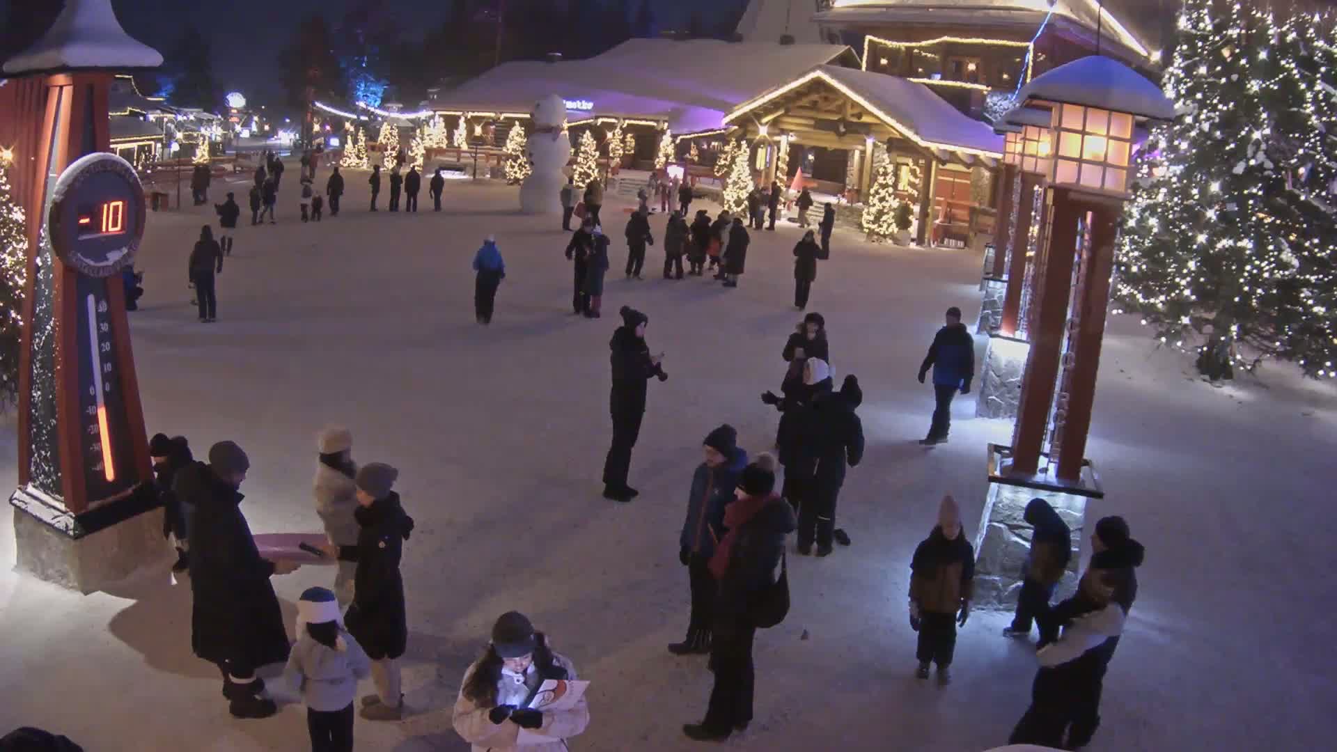 A snowy, nighttime scene shows many people gathered in a plaza, illuminated by Christmas lights and street lamps, with a large snowman visible in the background and a thermometer displaying -10 degrees.