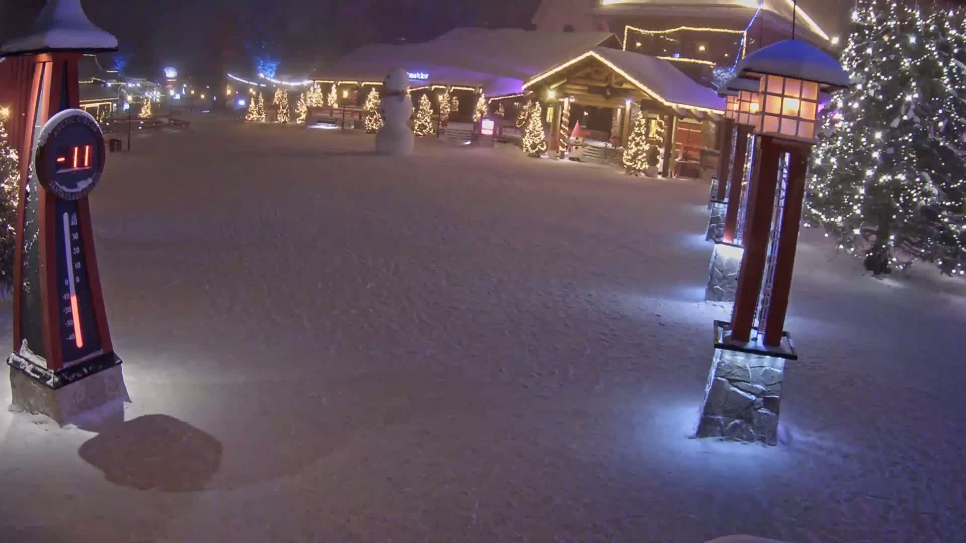 A snow-covered plaza at night, illuminated by lights on lampposts and Christmas trees, shows a thermometer indicating -11 degrees, with a snowman in the background.