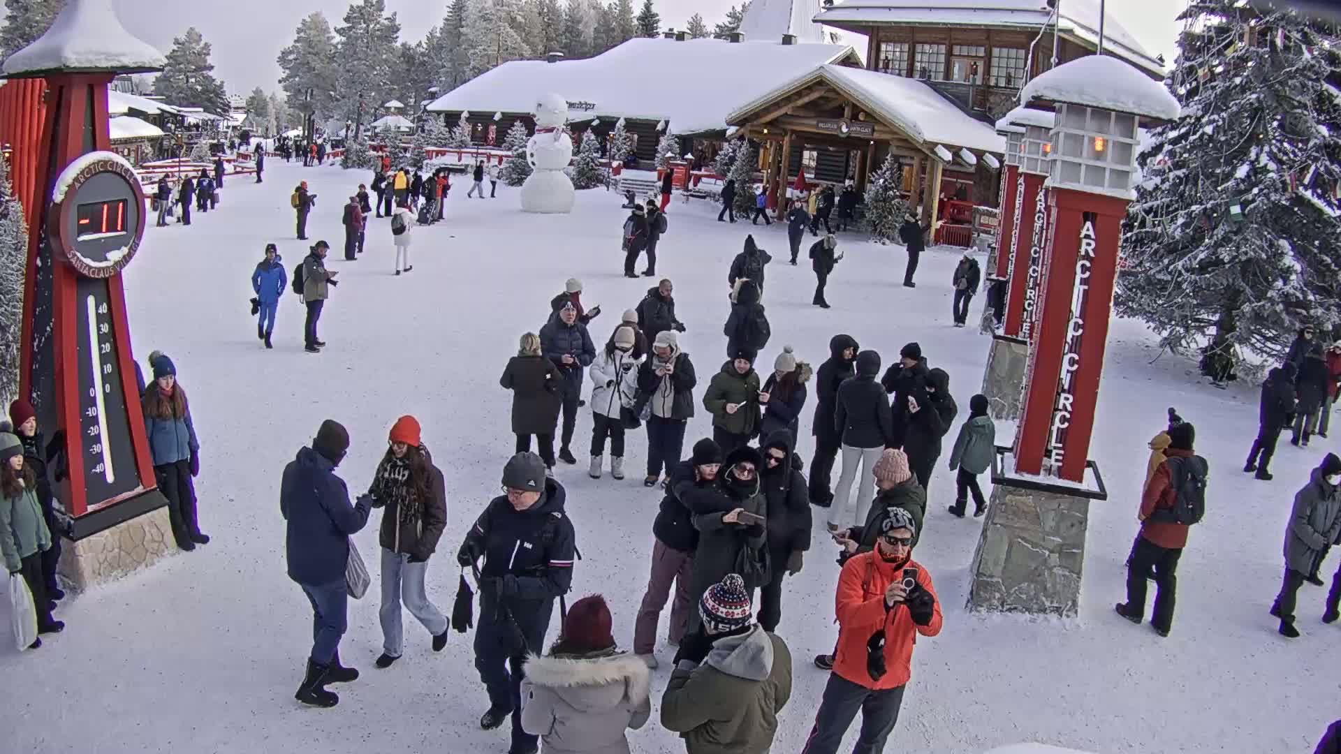 A crowd of people stands in a snowy plaza in front of wooden buildings, a large snowman is visible in the background.