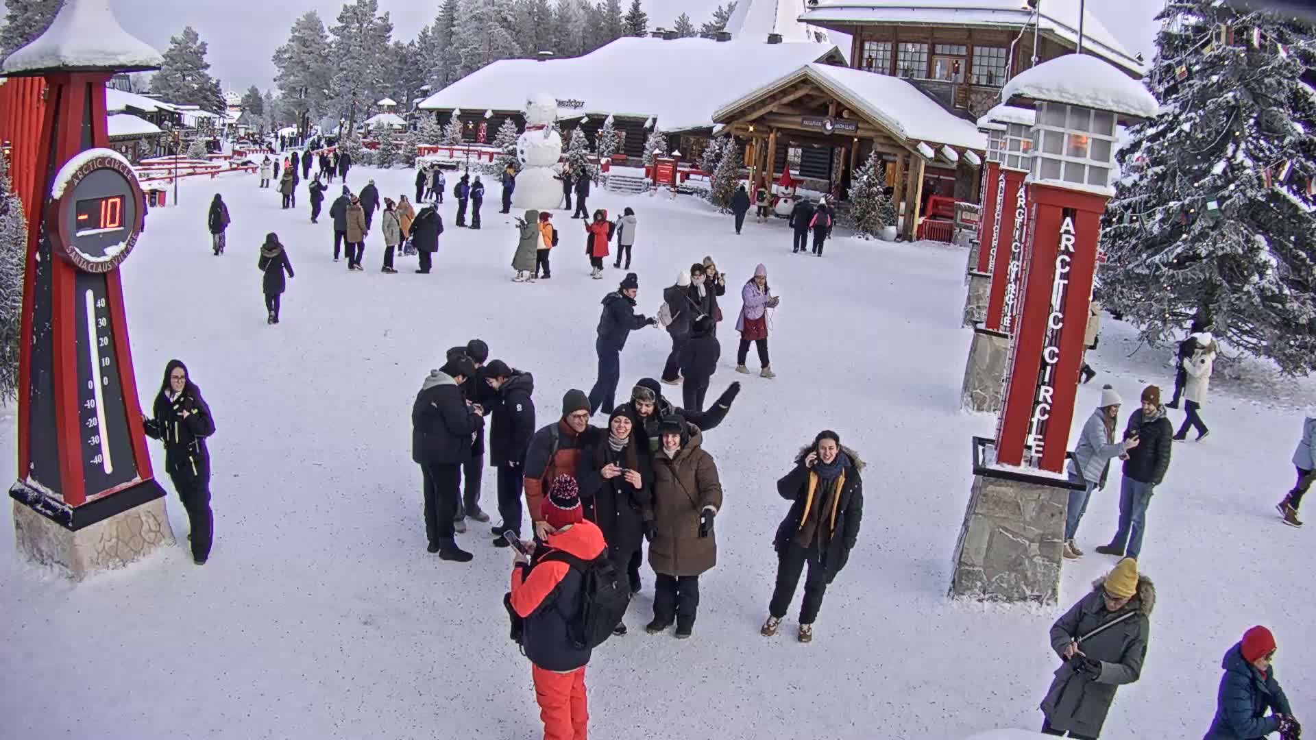 A snowy plaza with numerous people milling about, some in groups, near a building and a large snowman, with a thermometer showing -10 degrees.