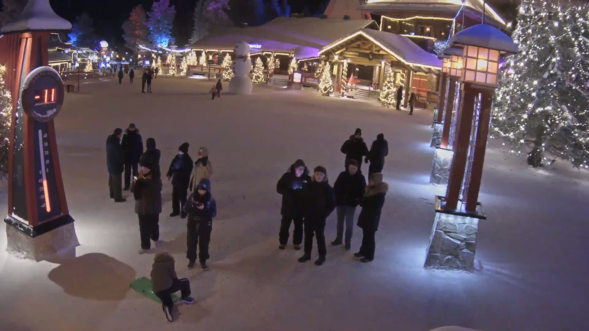 A snow-covered town square at night, illuminated by lights on buildings and lampposts, with several people gathered in groups and a child sledding, and a large snowman visible in the background.