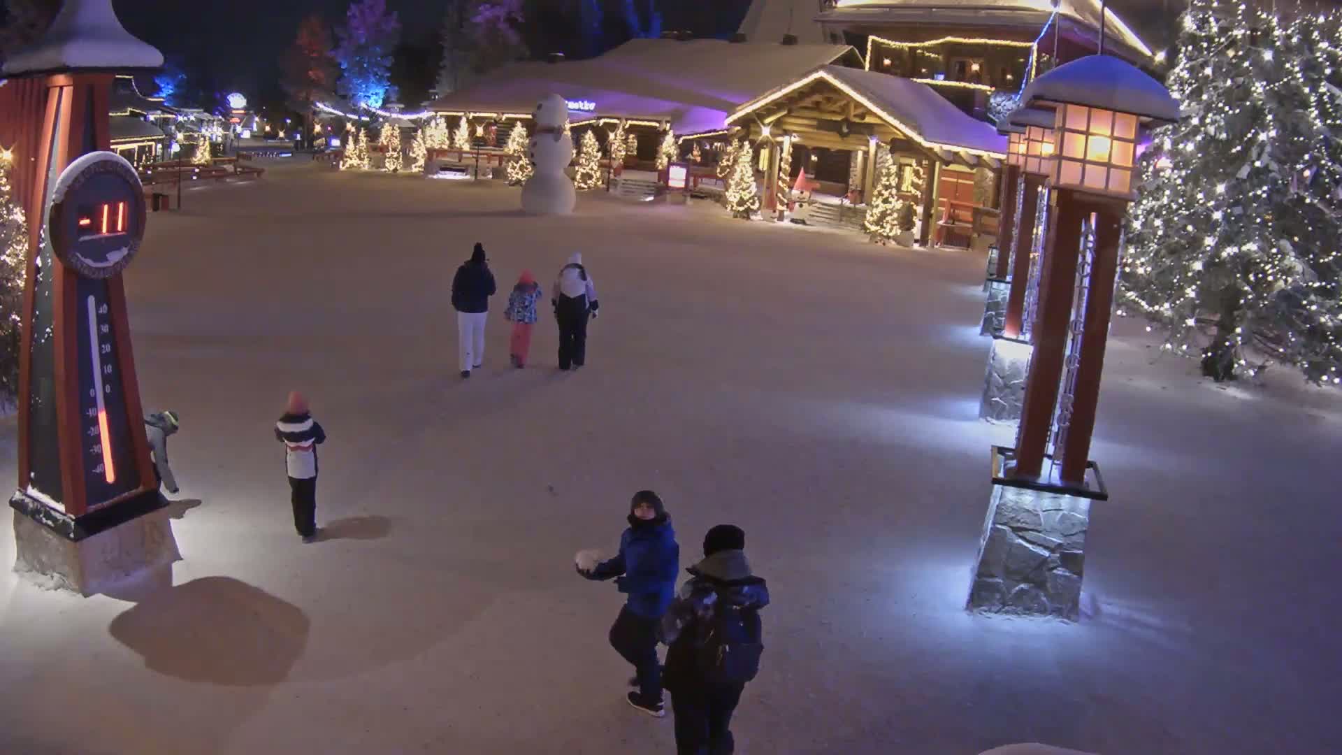 A snow-covered plaza at night, illuminated by lights, features several people walking, a large snowman, and a thermometer showing -11 degrees.