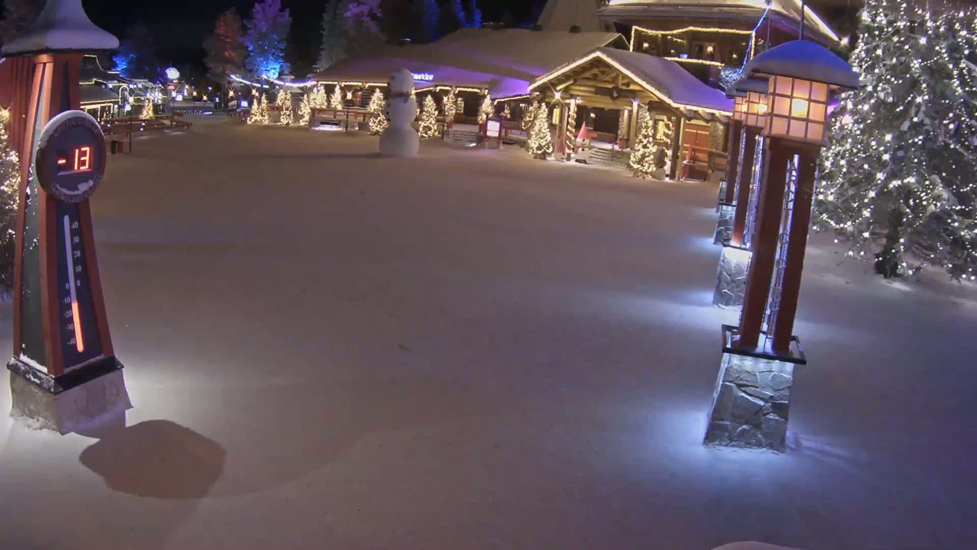 A snow-covered village square at night shows a snowman near a building with Christmas lights, illuminated light posts, and a thermometer displaying -13 degrees.