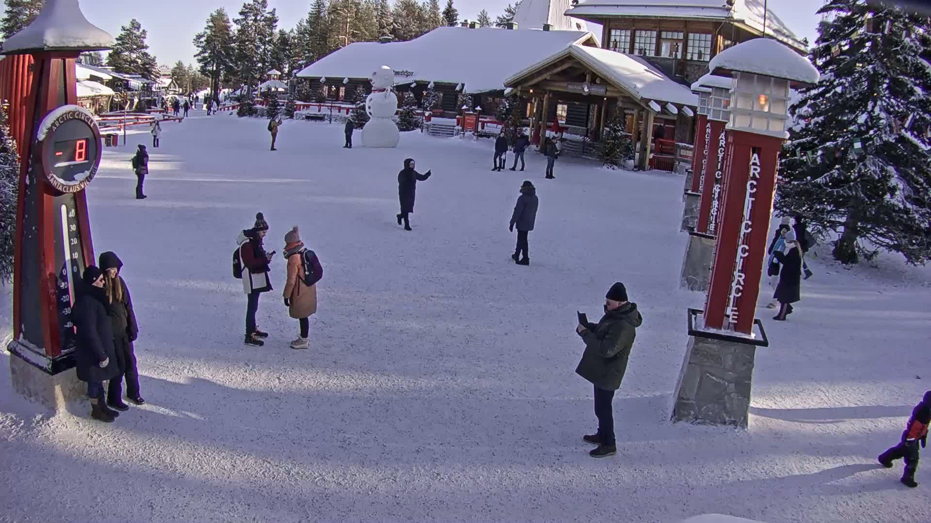 A snowy, sunny scene shows several people walking around a village square, with a large snowman and buildings in the background.