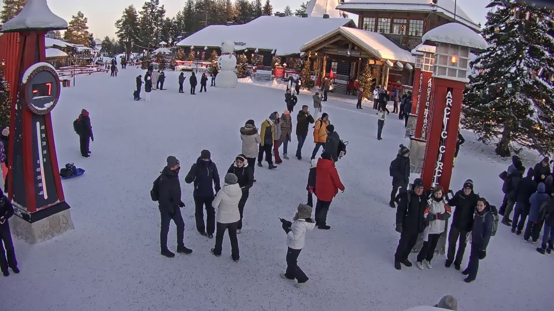 A snowy plaza, populated by numerous people, features a snowman, wooden buildings, and tall red signposts.